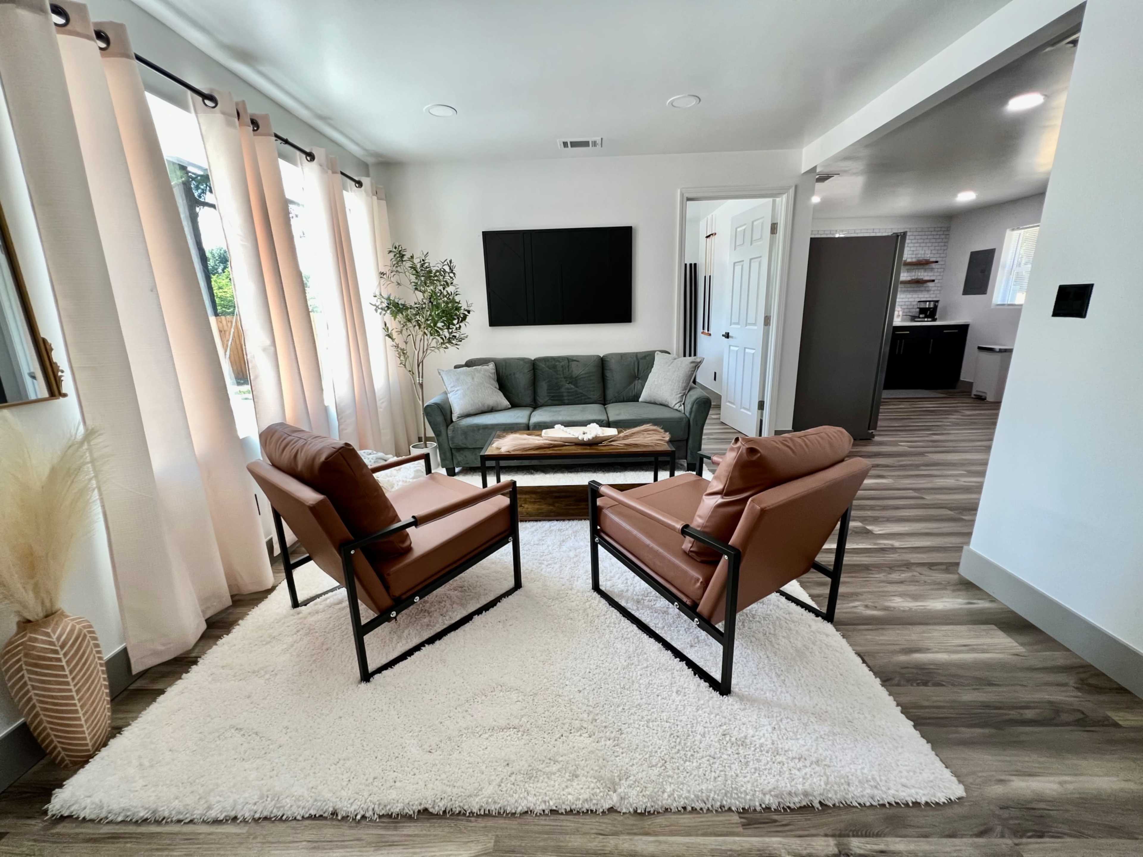 A modern living room with a gray sofa, two brown chairs, a coffee table, and large windows dressed with light curtains.