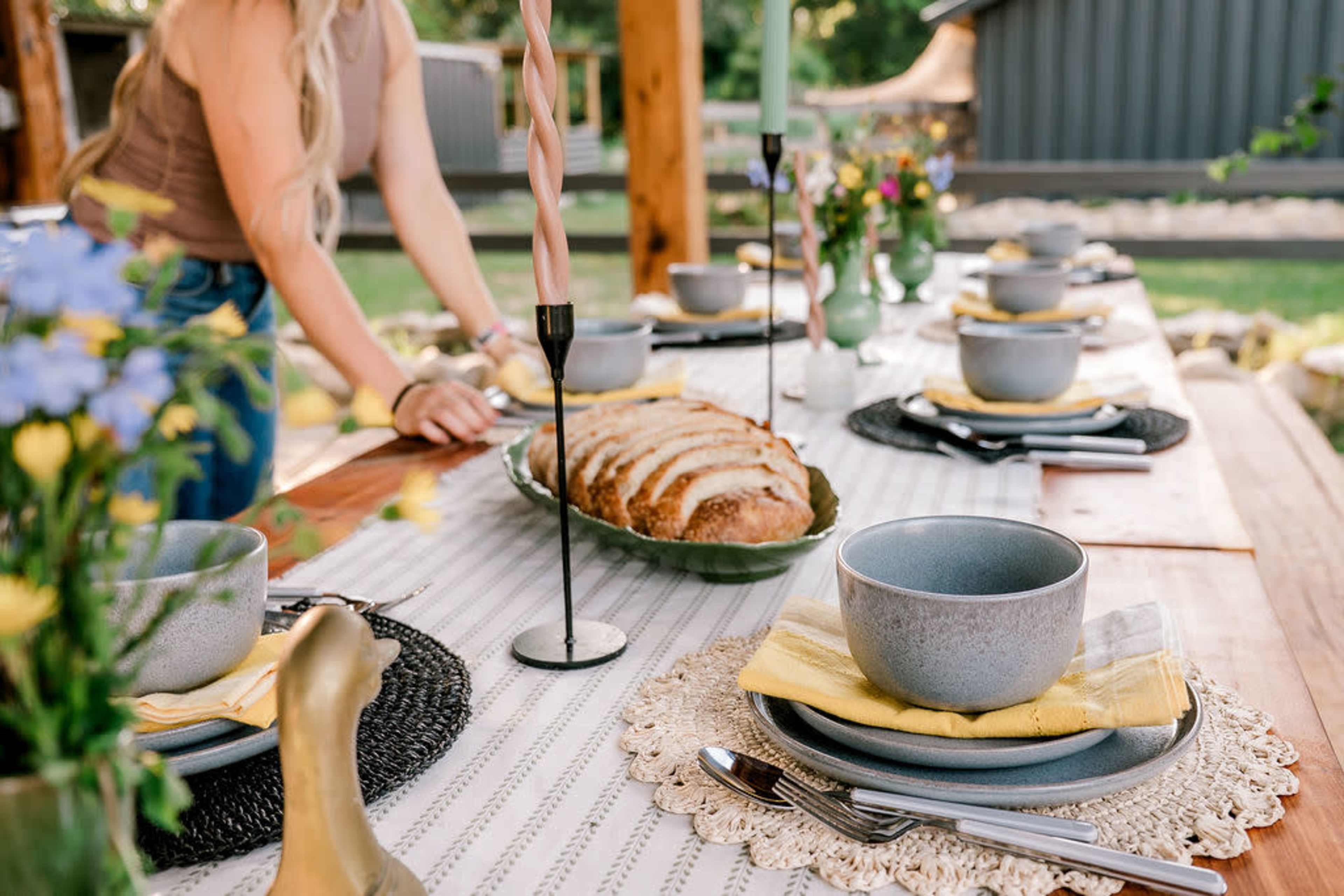 A long wooden table is set with bowls, plates, candles, and a loaf of bread, while a person arranges the table decorations in a garden setting.