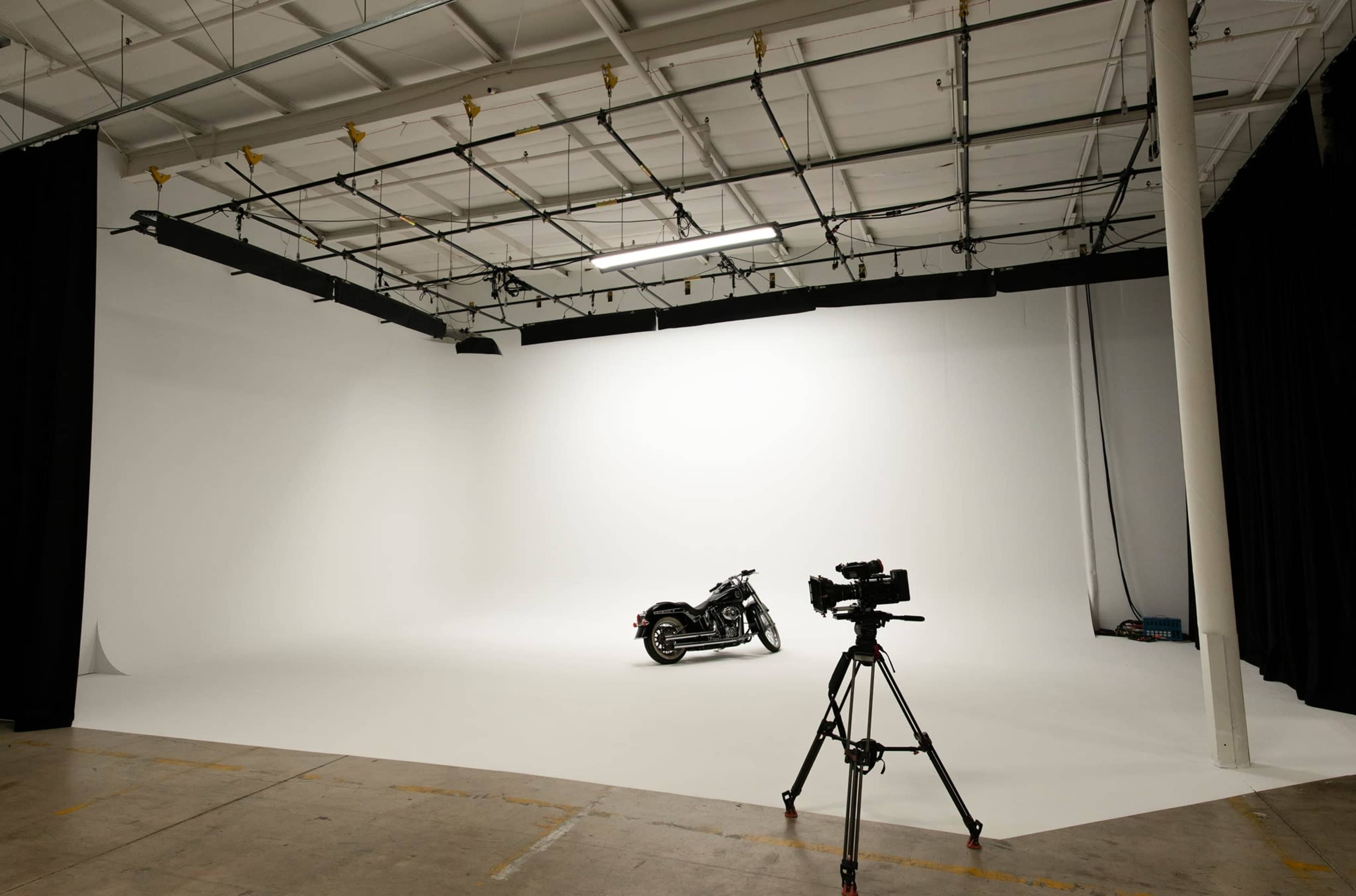 A motorcycle is positioned on a white stage with a camera set up to film it in an empty studio space.