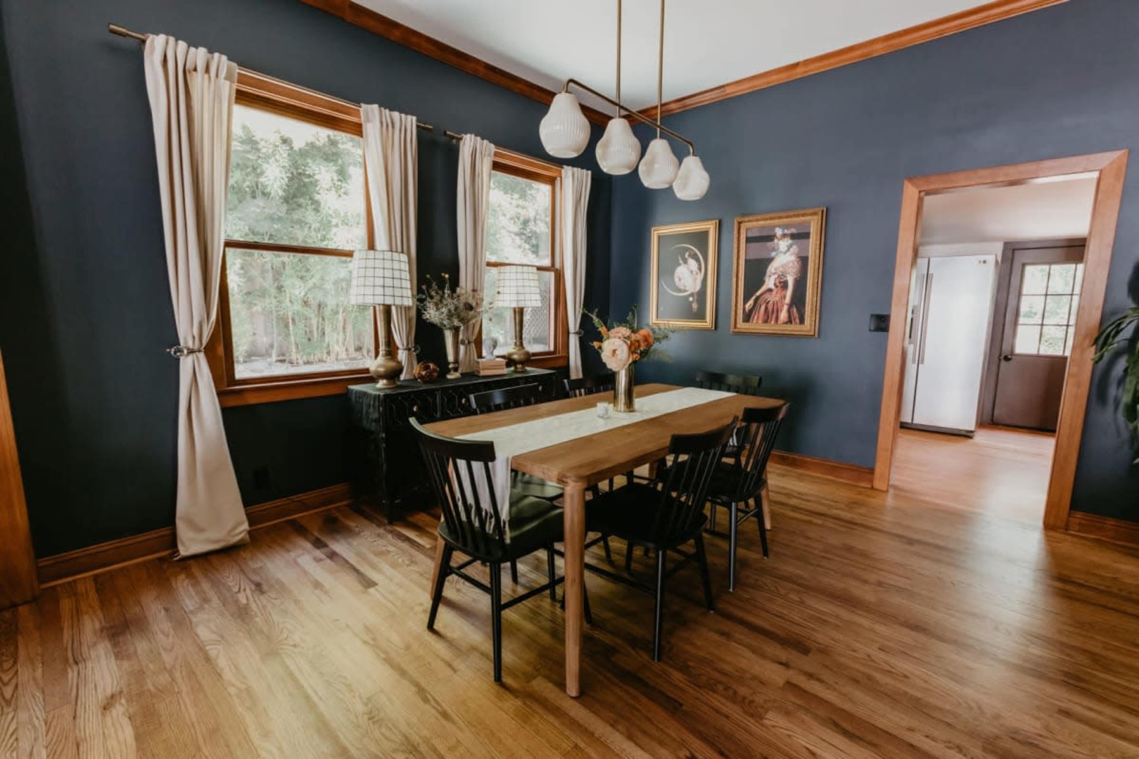 The image shows a dining room featuring a wooden table with six black chairs, framed artwork on the walls, and large windows with curtains.