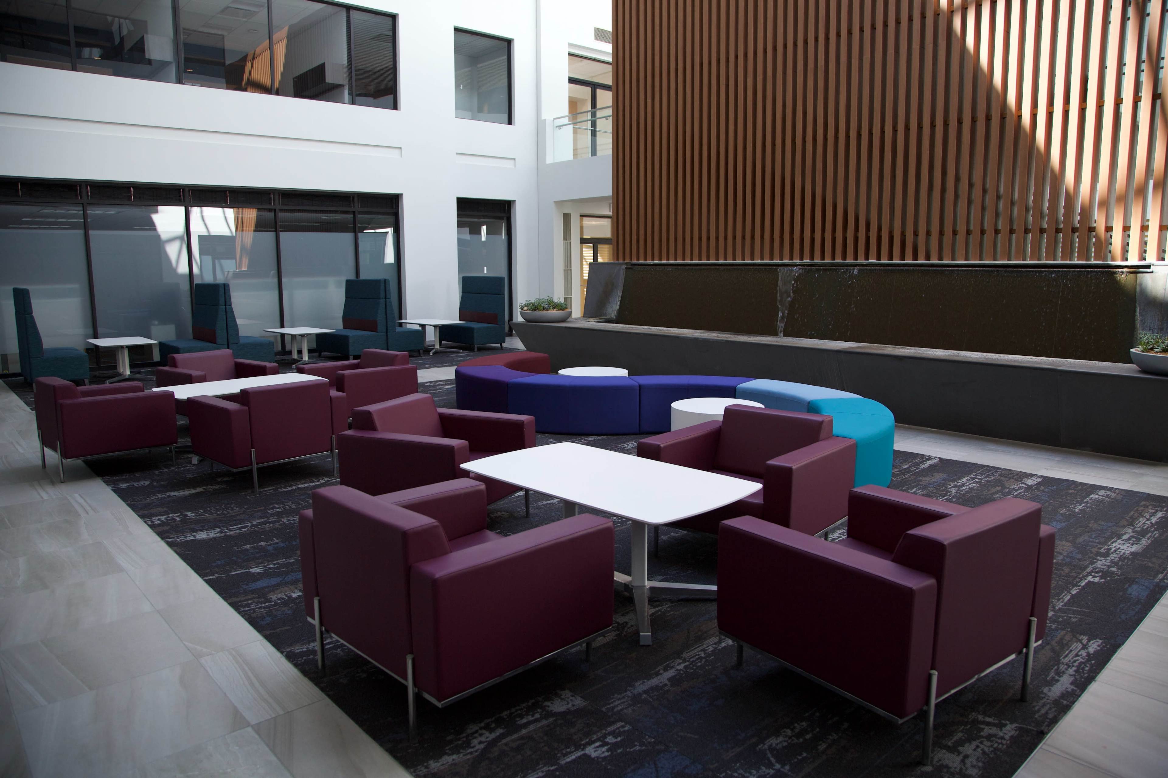 The image shows a modern indoor seating area with purple and blue chairs arranged around white tables, near a water feature.
