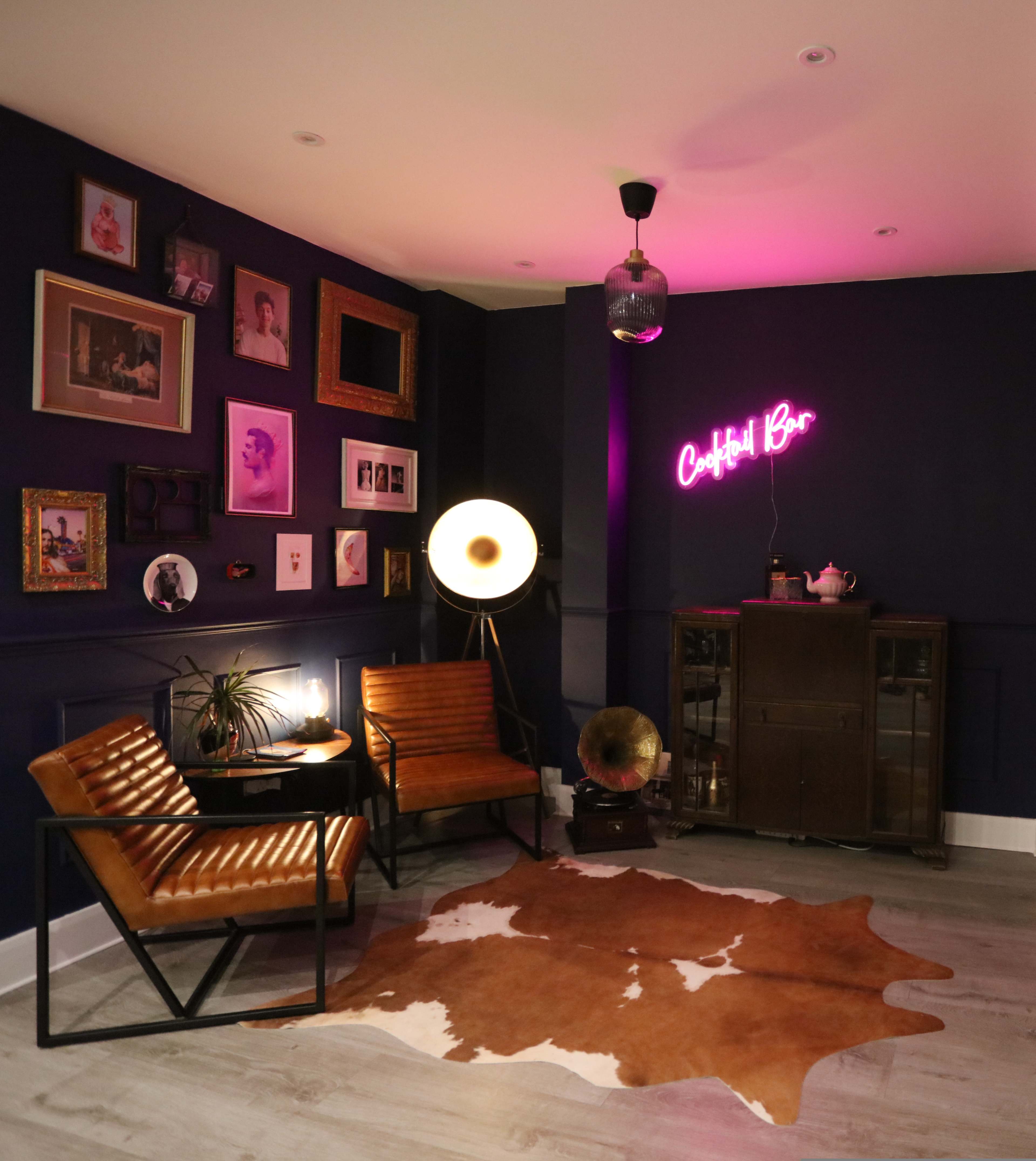The image shows a stylish corner of a room with dark purple walls, featuring framed artwork, a neon "Cocktail Bar" sign, a vintage gramophone, and mid-century modern furniture.