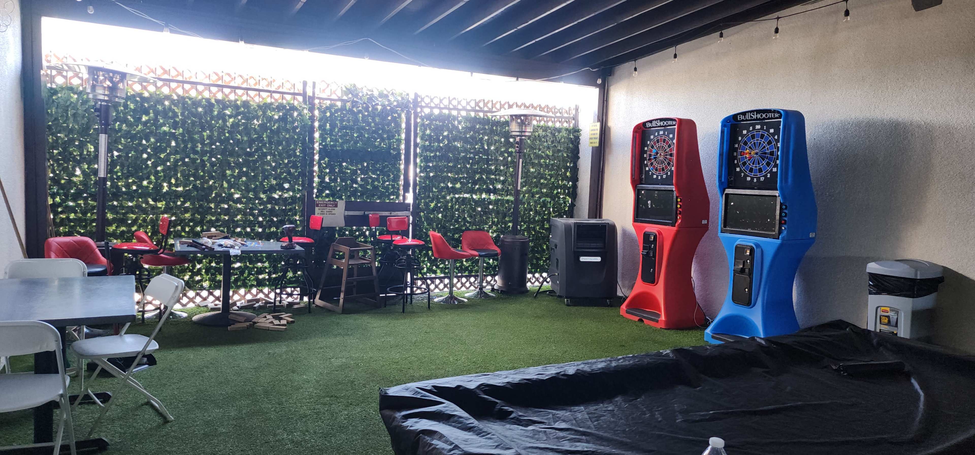 The image shows a recreational area featuring two dartboards, several chairs and tables, and artificial grass flooring under a covered patio.