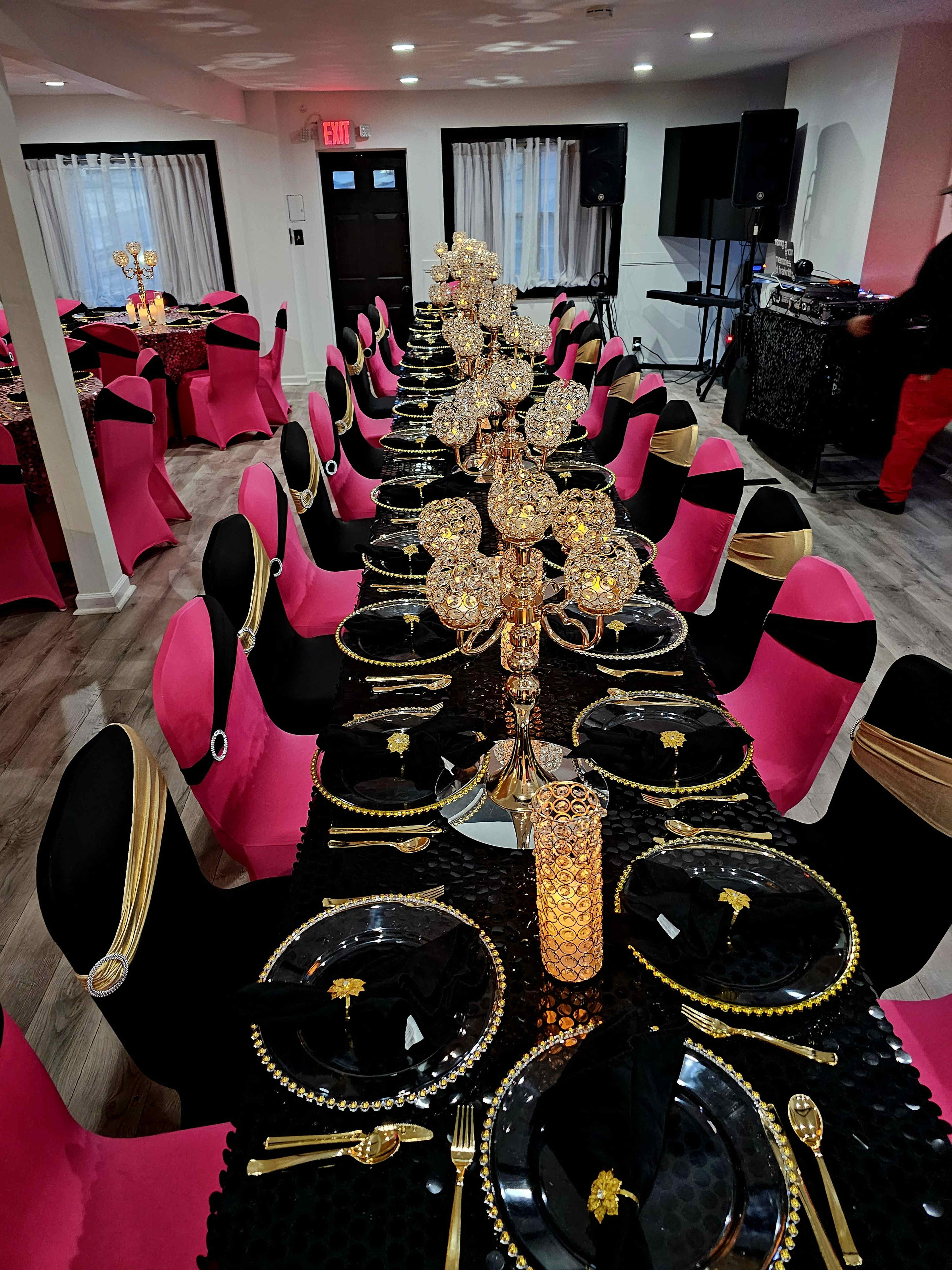 A long banquet table is set with black plates, gold utensils, and ornate candle holders, surrounded by pink and black chairs in a decorated event space.