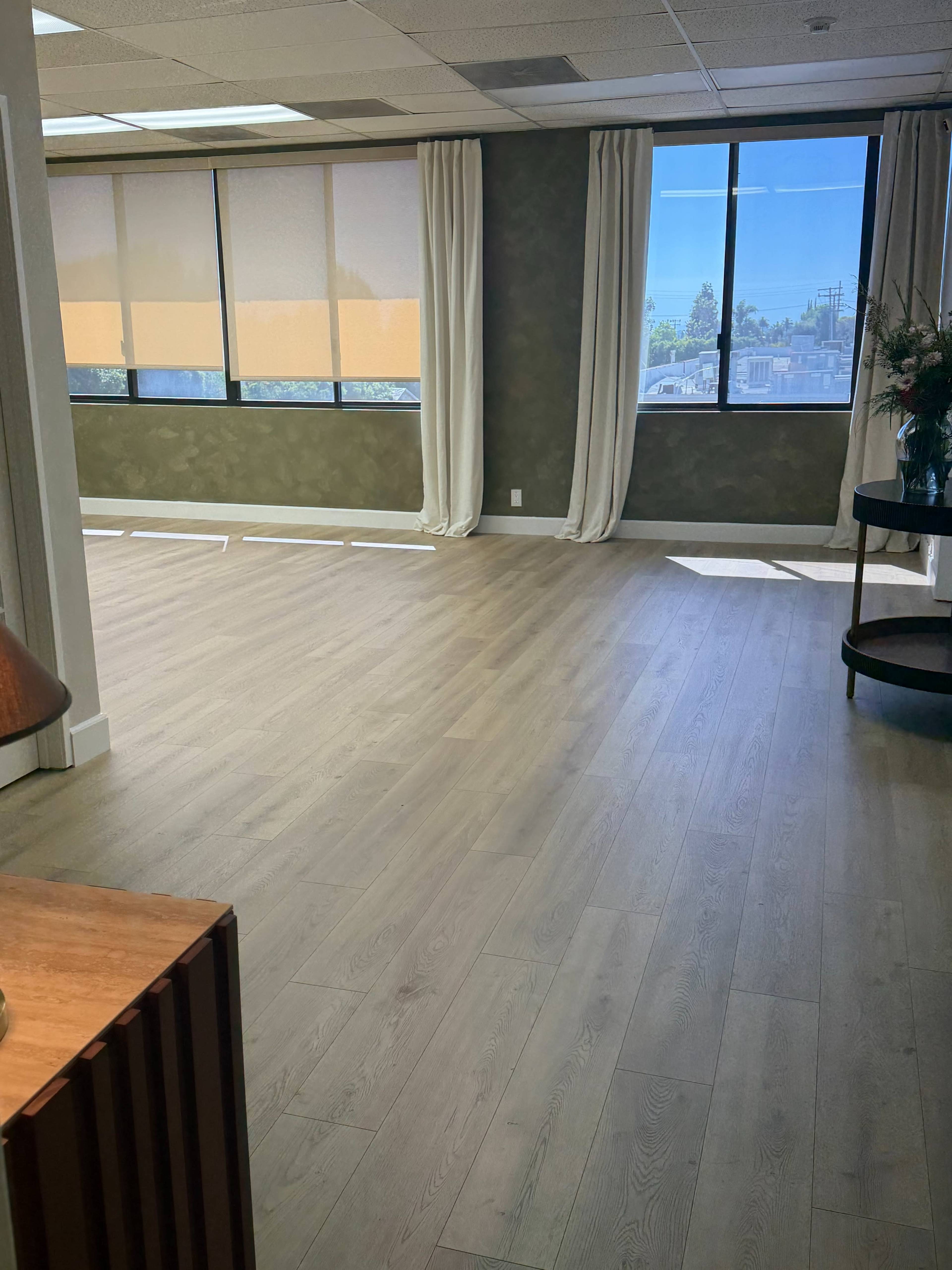 The image shows a spacious, well-lit room with wooden flooring, large windows featuring sheer curtains, and a small round table in the corner.