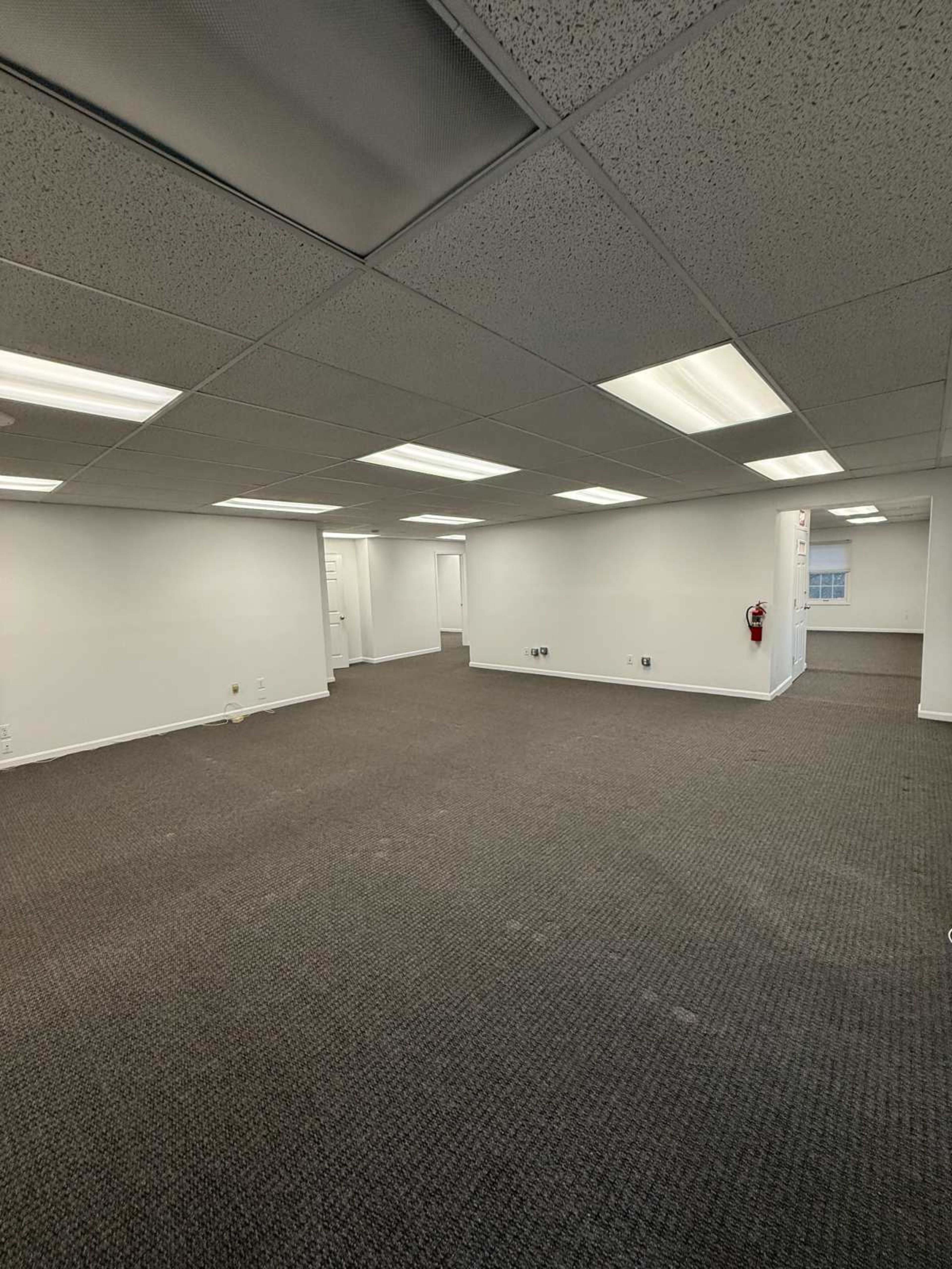 The image shows a spacious, empty office with ceiling lights, carpeted flooring, and white walls.