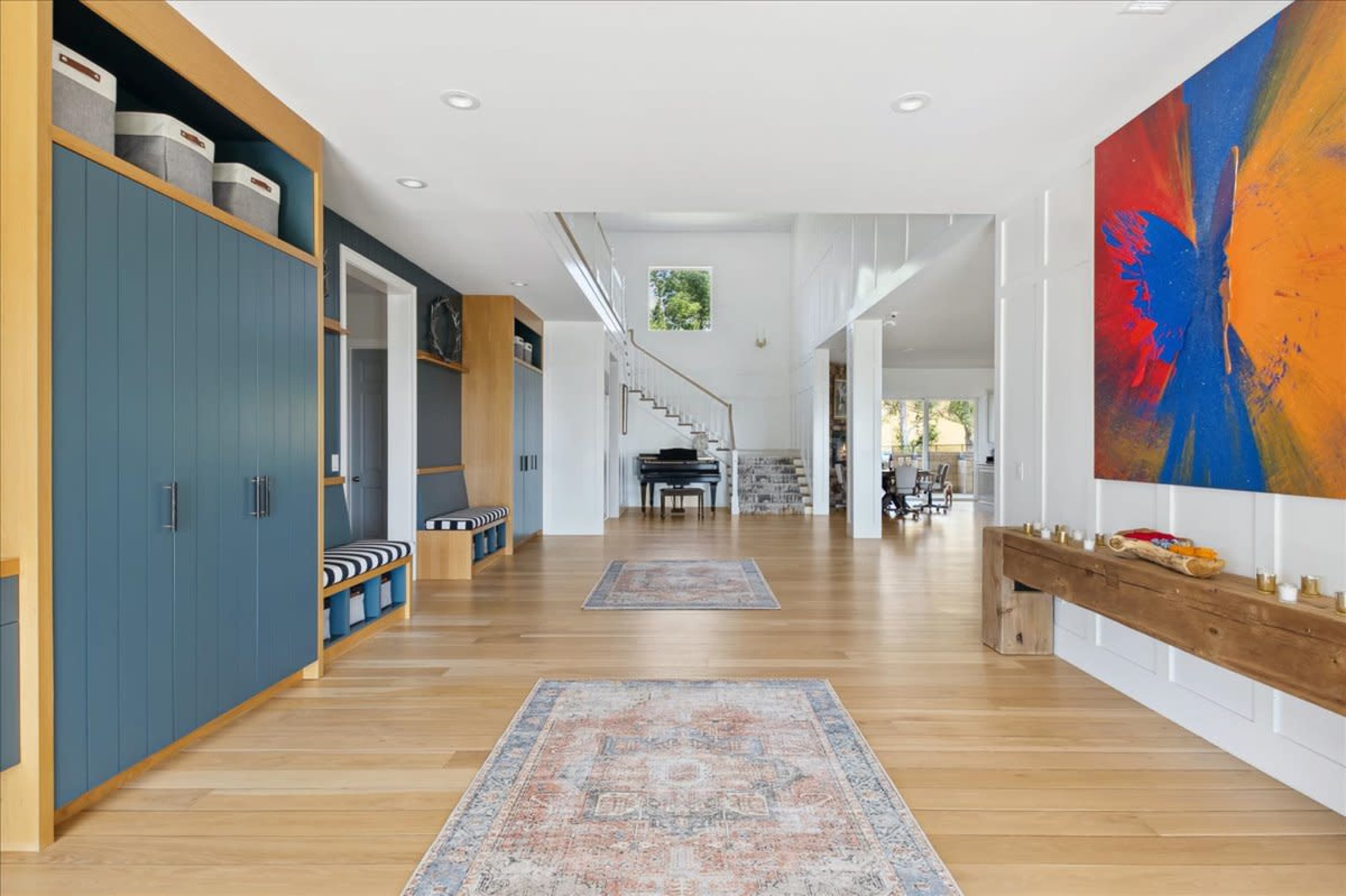 The image shows a spacious hallway with wooden flooring, built-in storage cabinets, decorative rugs, and an abstract painting on the wall.