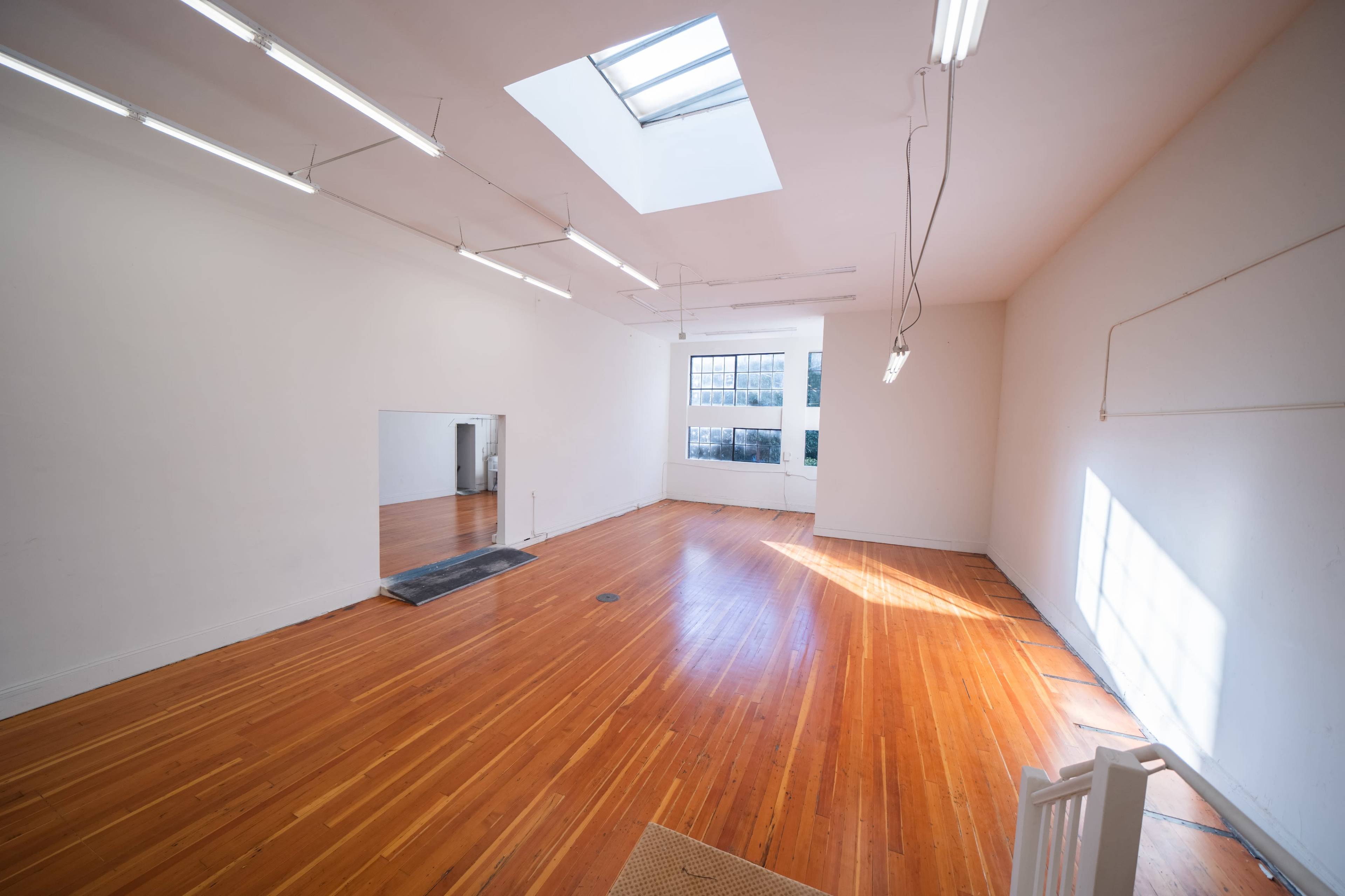A spacious, empty room with hardwood floors, a skylight, and a large window, illuminated by fluorescent lights.