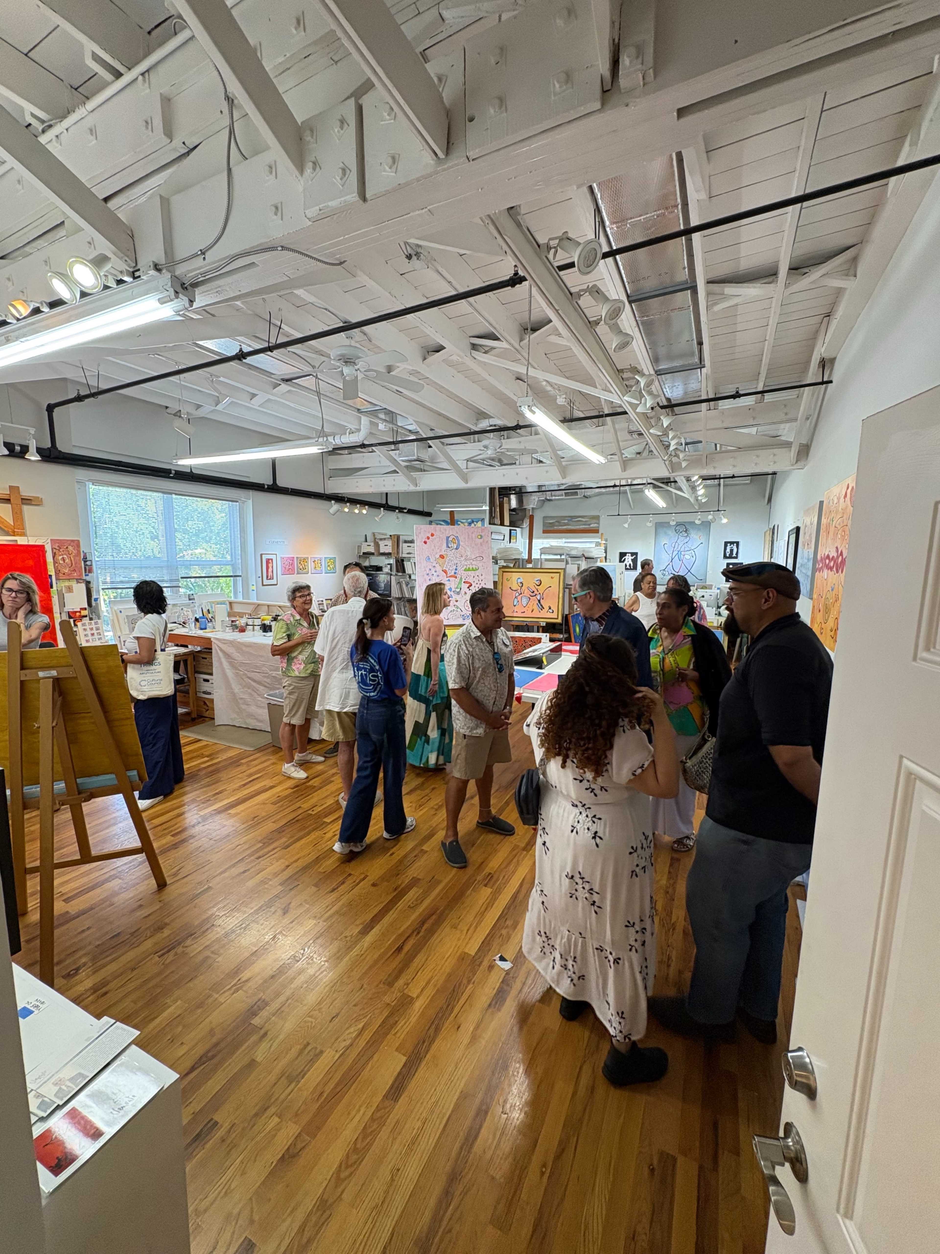A group of people is viewing artwork in a bright, open gallery space with wooden floors and white walls.