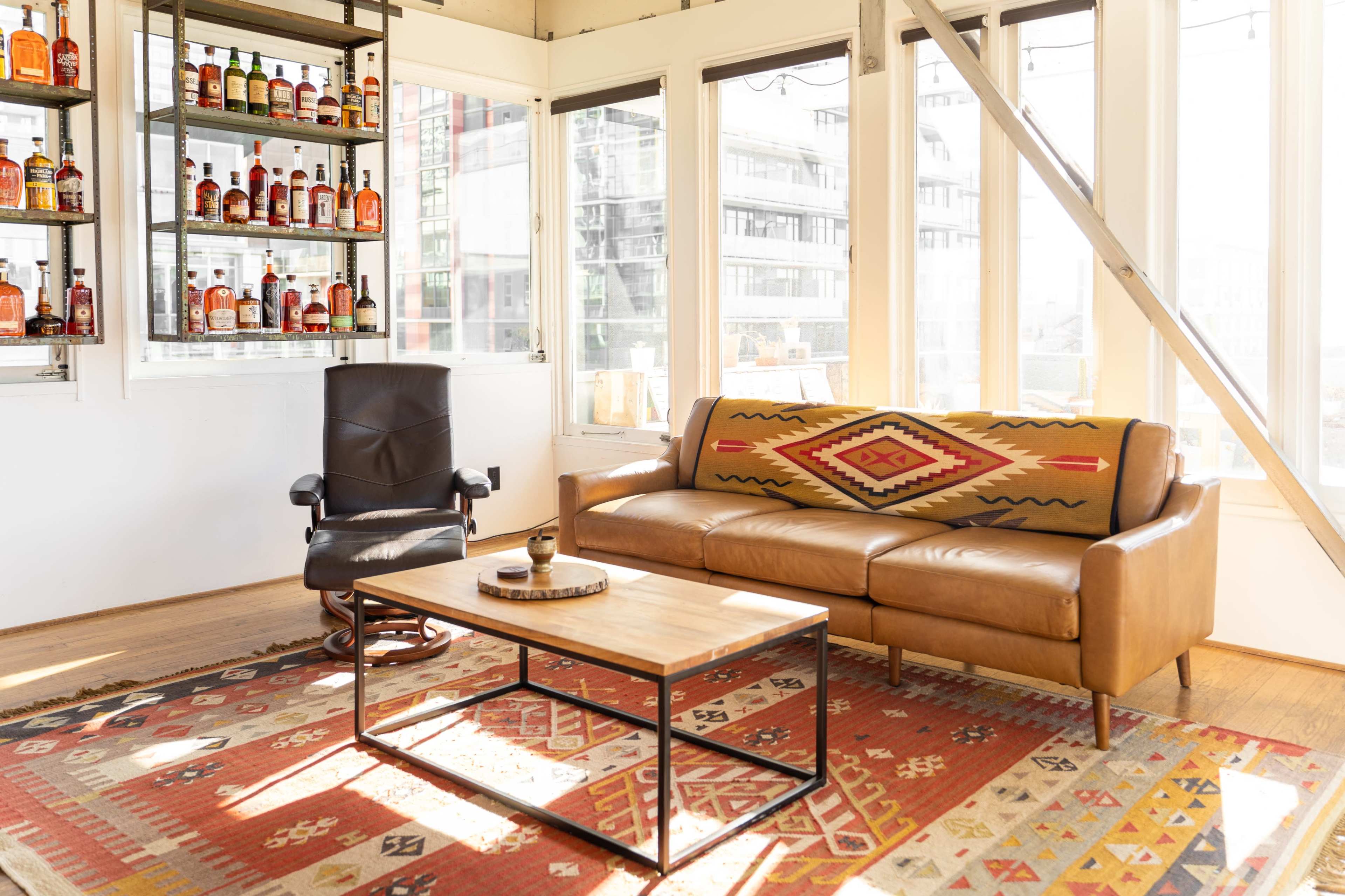 A modern living room features a brown leather sofa with a patterned throw, a wooden coffee table, and a black rocking chair, surrounded by large windows and a shelf displaying various bottles.