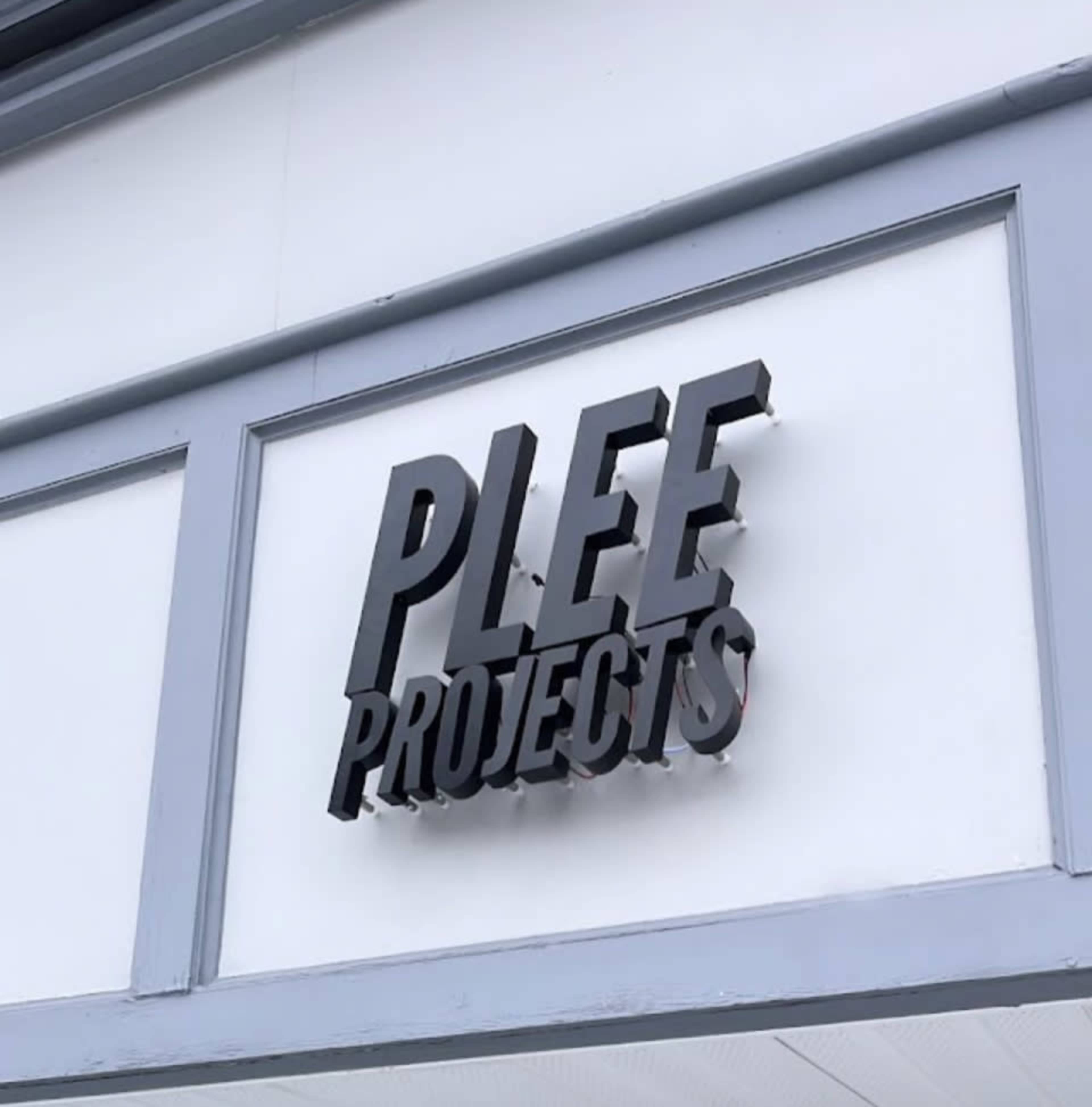 A three-dimensional sign displaying "PLEE PROJECTS" is mounted on a white wall.