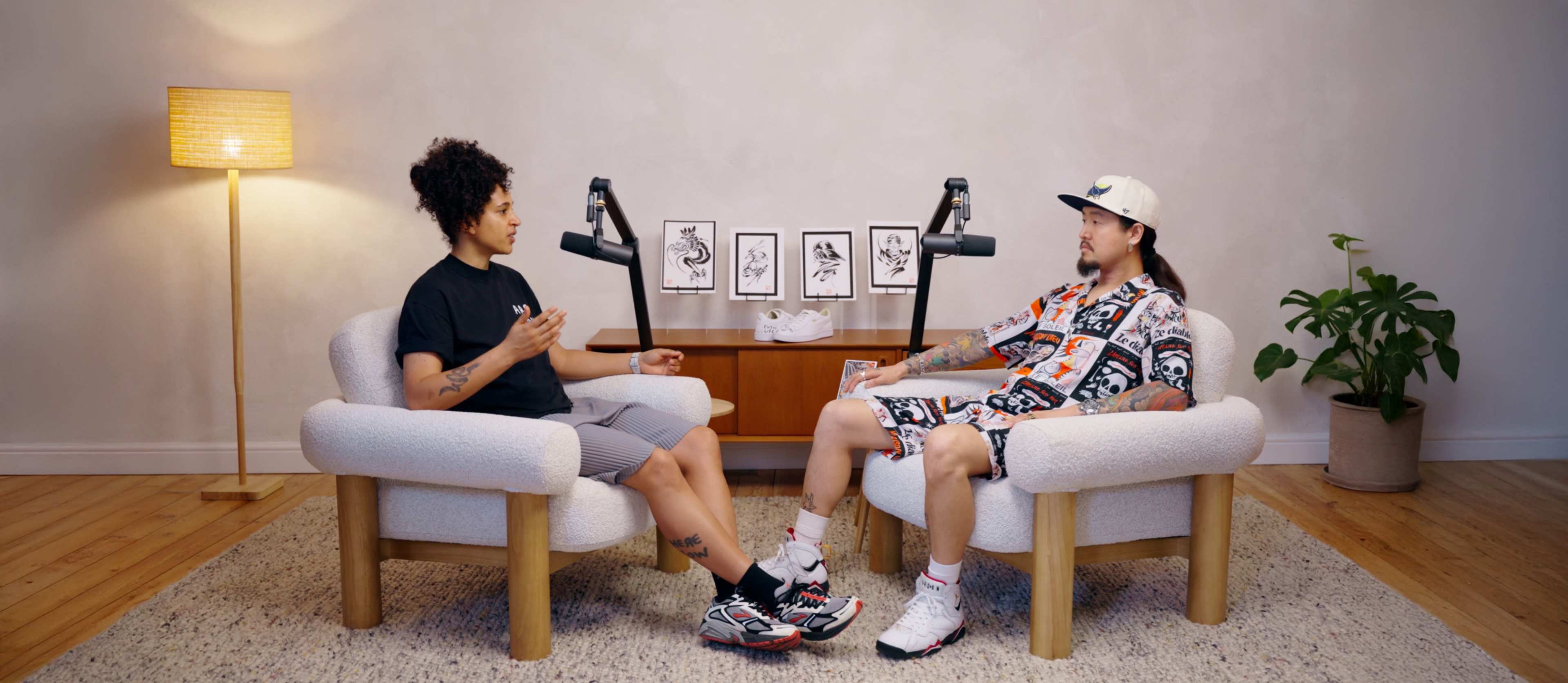 Two people are seated in modern armchairs facing each other, engaged in conversation, with framed artwork on a wooden shelf in the background.