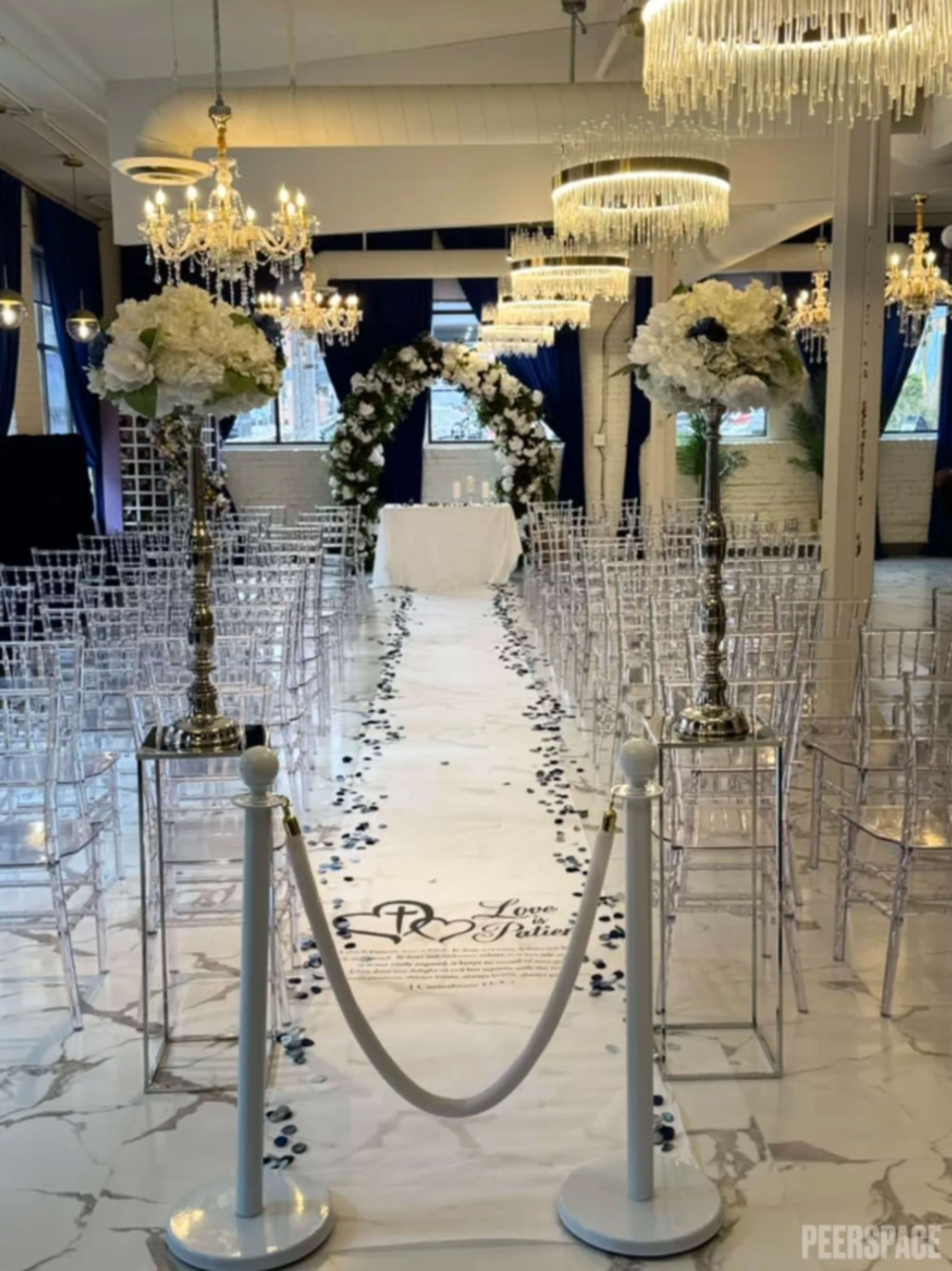 The image shows a wedding venue decorated with white flowers, clear chairs arranged in rows, and an altar at the end of a pathway adorned with rose petals and a white carpet.