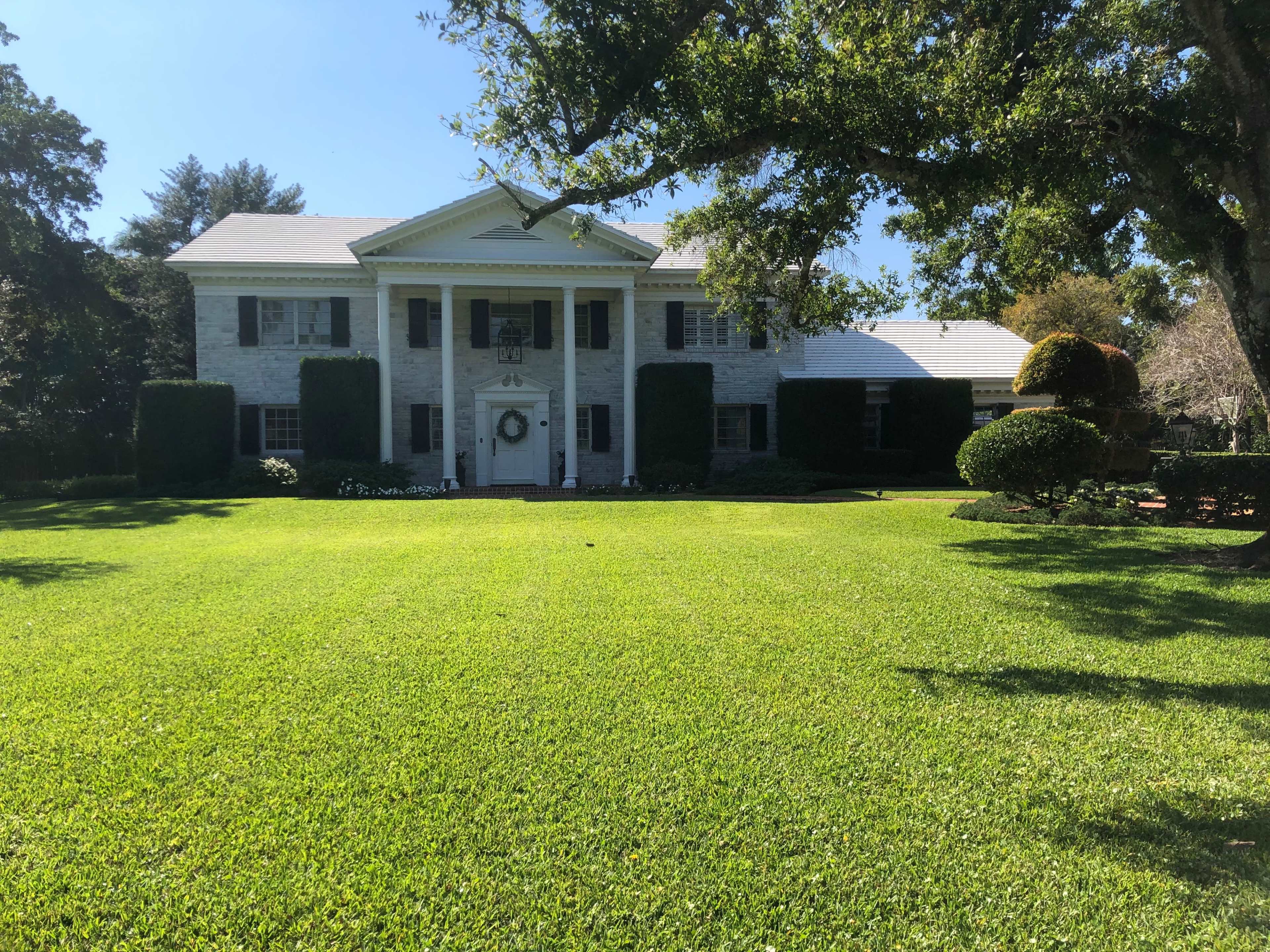 A large, two-story house with white stone exterior and black shutters sits on a well-manicured lawn surrounded by neatly trimmed bushes and trees.