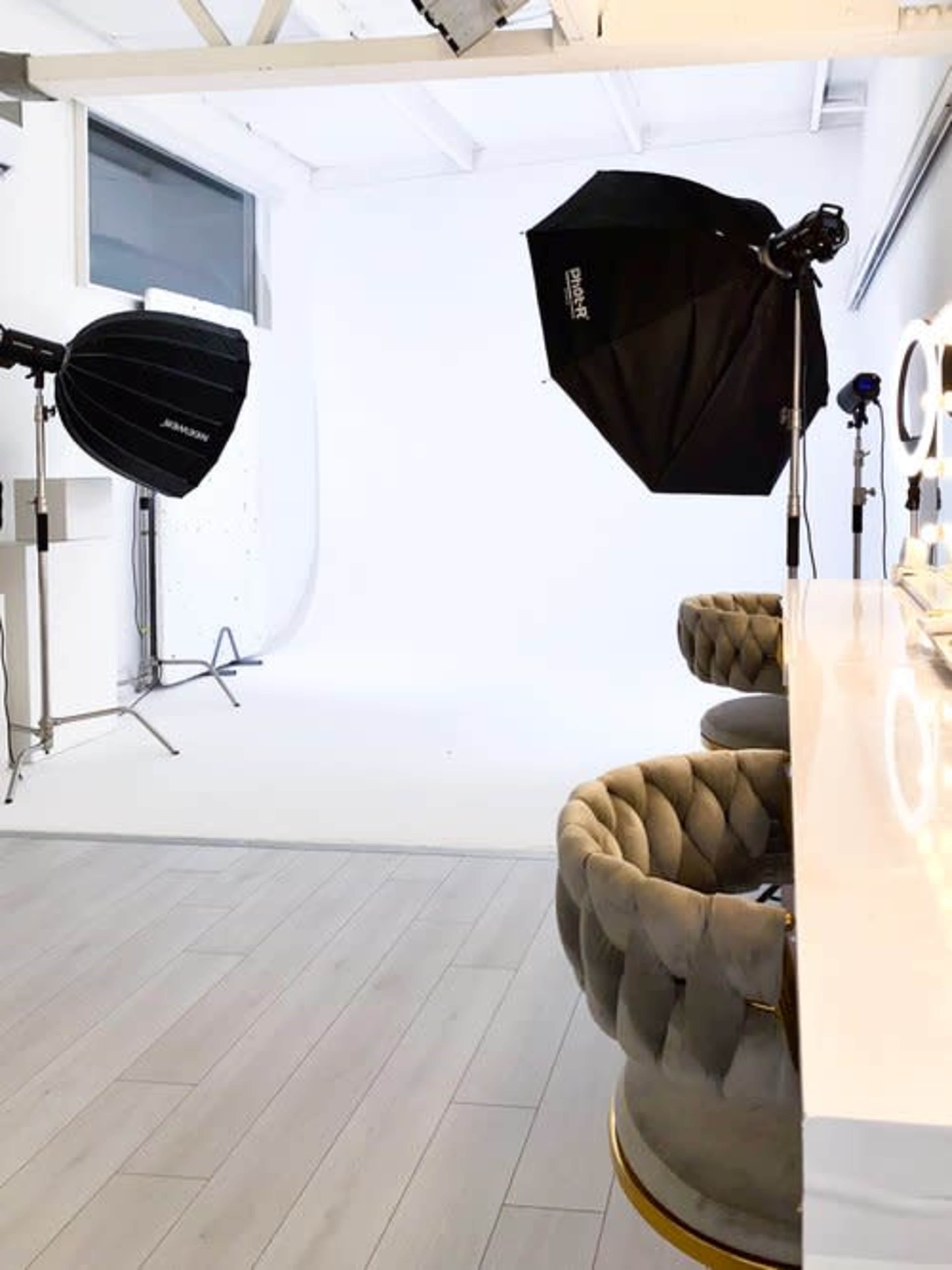 The image shows a photography studio with two softbox lights and a pair of tufted chairs set against a white backdrop.