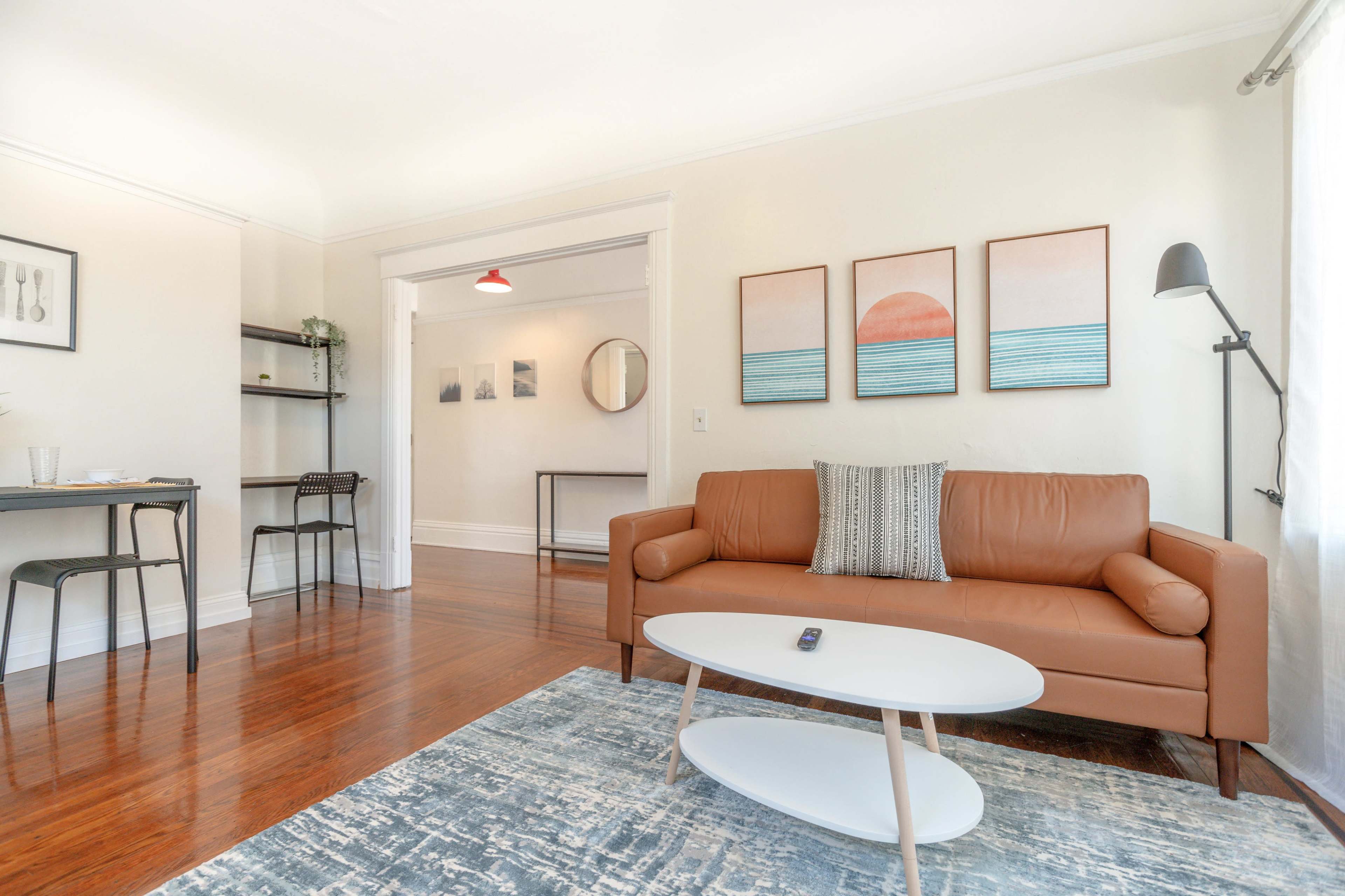 A living room featuring a brown leather sofa, a round coffee table, and framed wall art above the couch, with a dining area visible in the background.