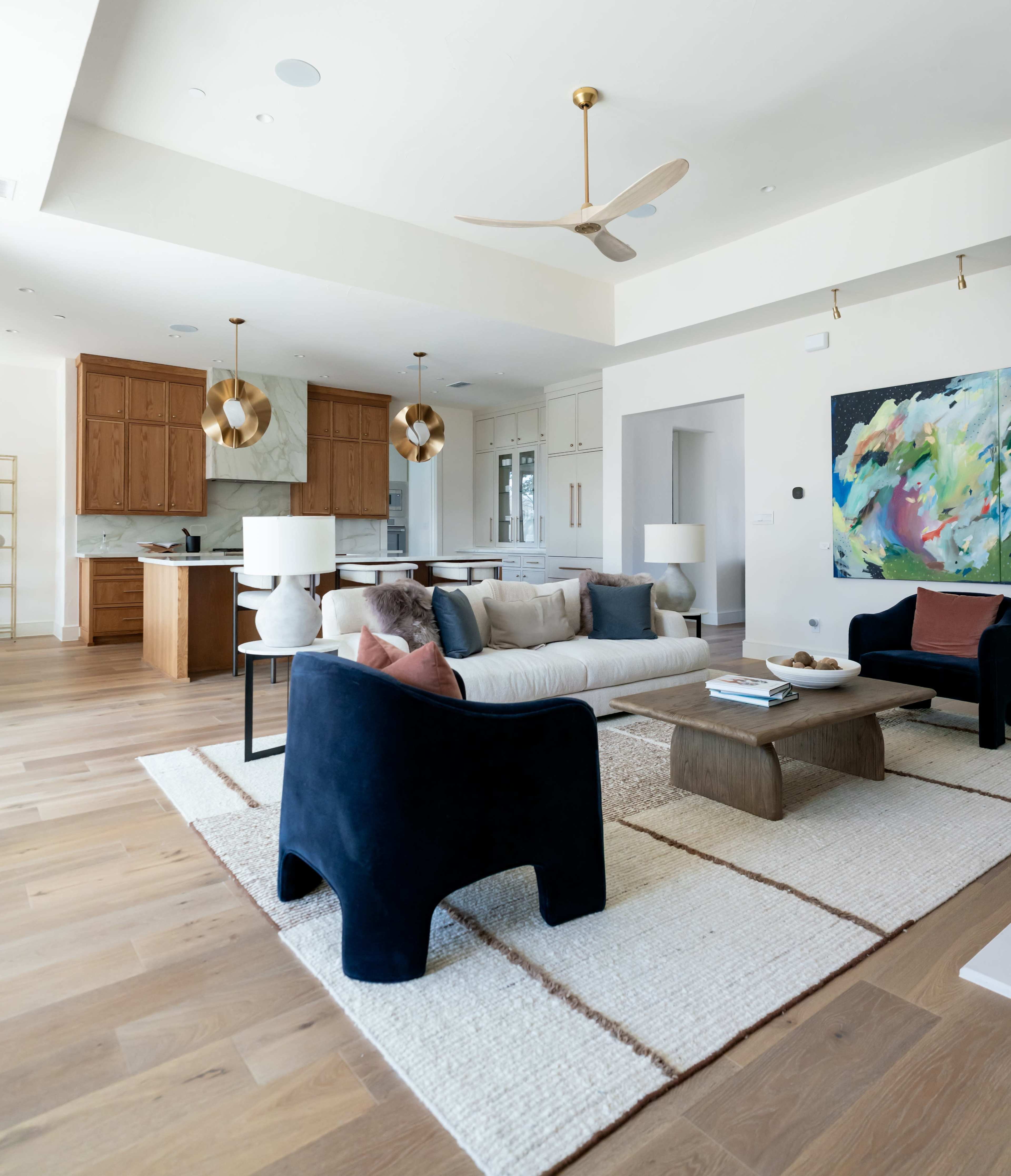 The image depicts a modern living room with a beige sofa, two navy blue armchairs, and a wooden coffee table, adjacent to a kitchen with wooden cabinets and pendant lighting.