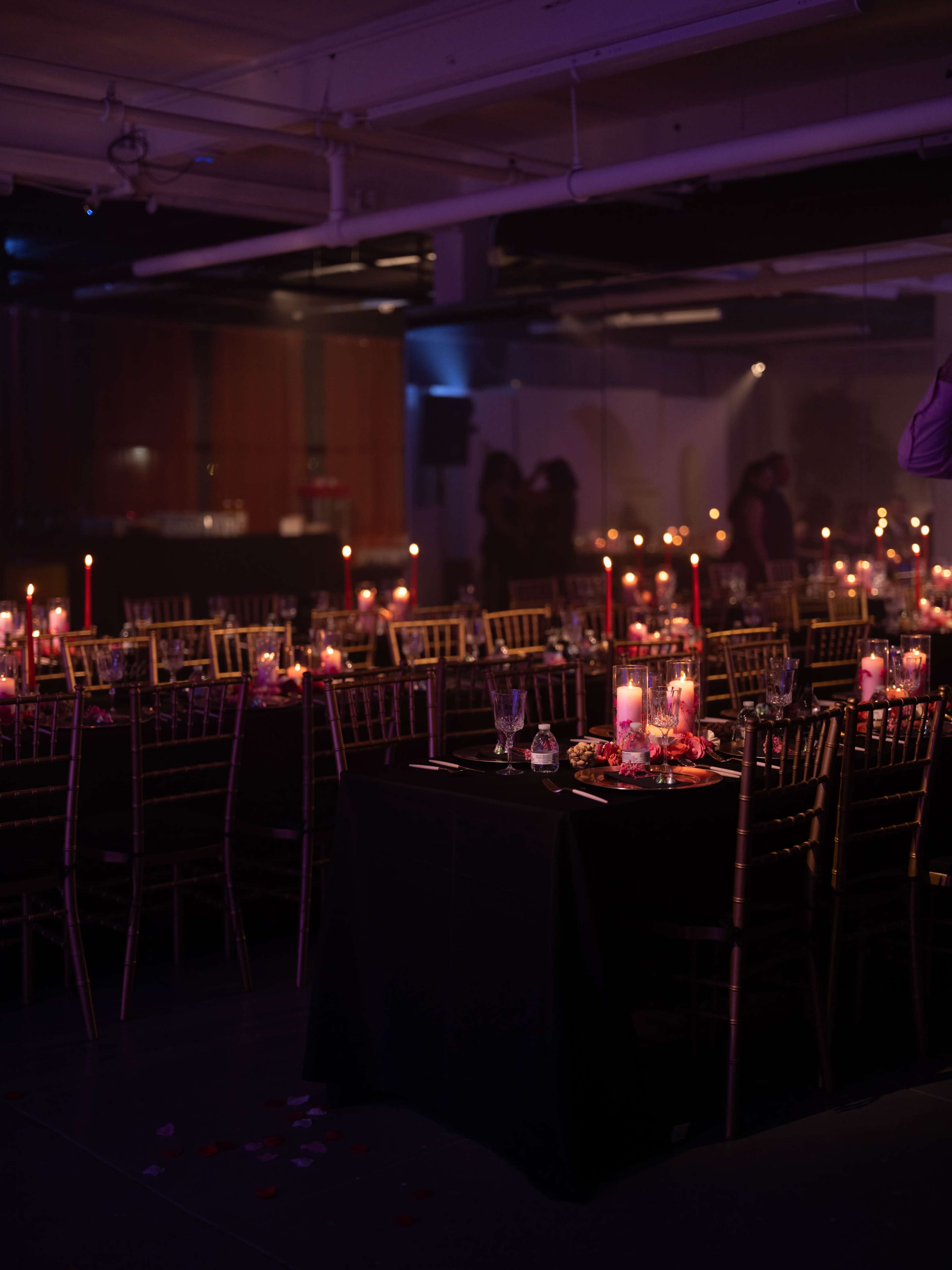A dimly lit event space is set up with multiple tables adorned in black linens, gold chairs, and candles, creating an elegant atmosphere.