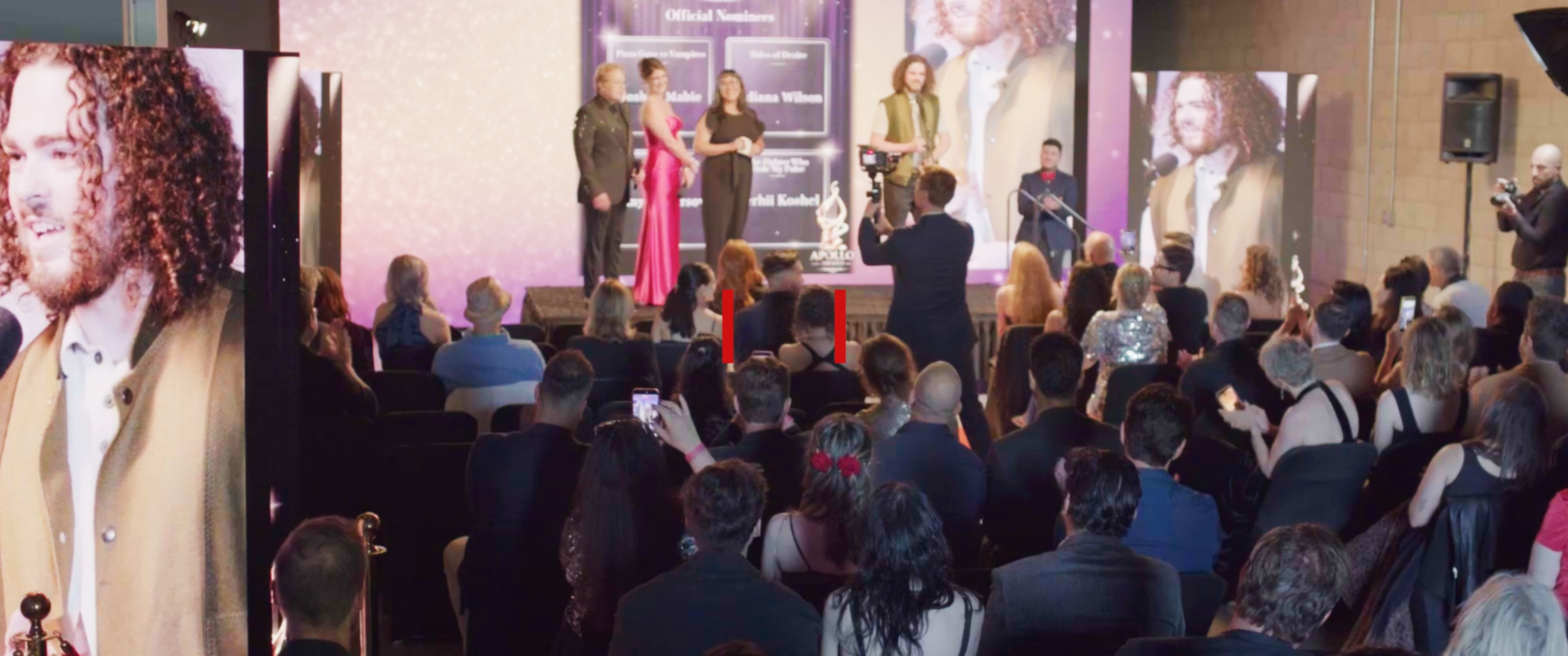 A crowd gathers at an awards ceremony, where presenters are on stage acknowledging a recipient as attendees capture the moment with their phones.