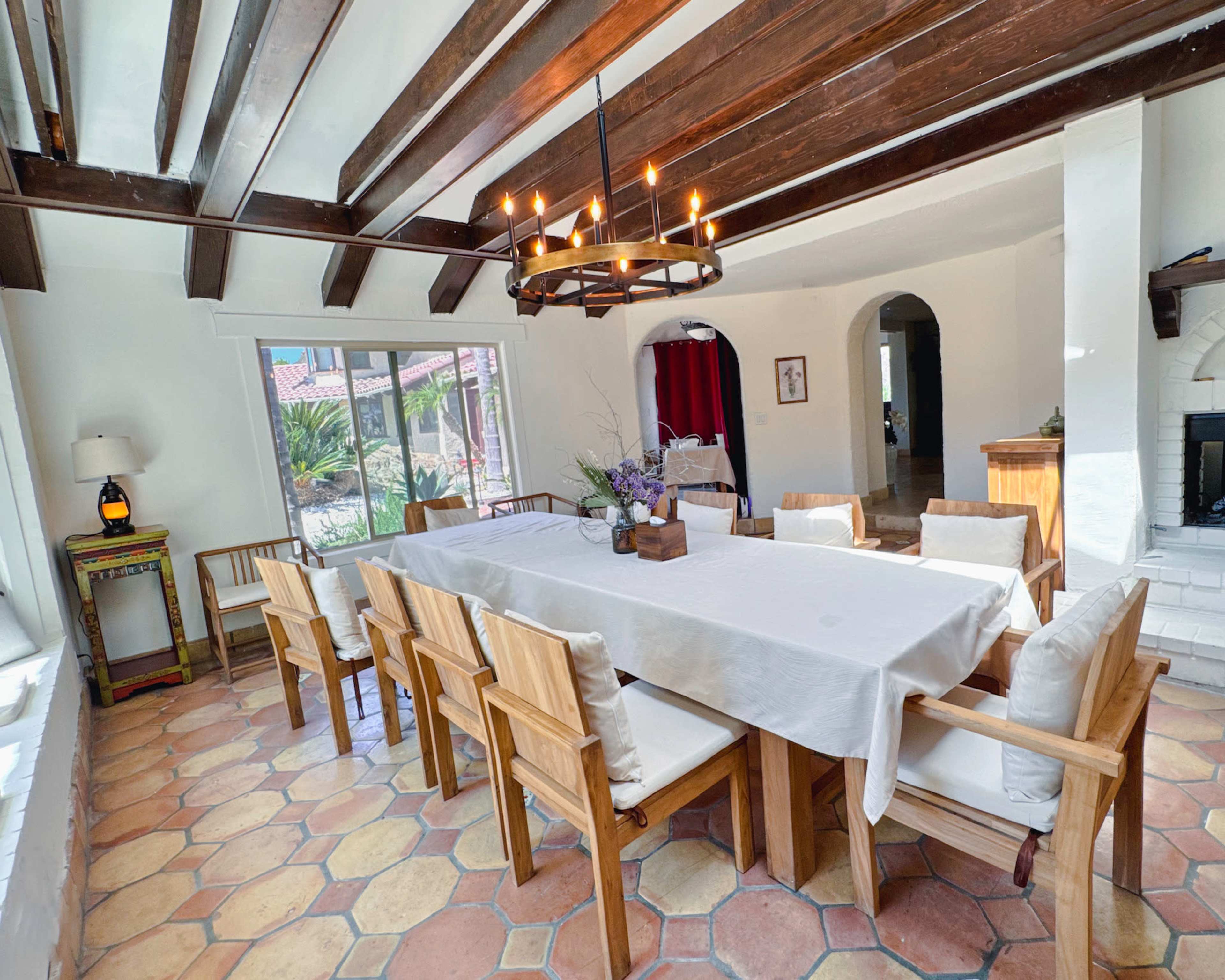 A spacious dining room features a large wooden table surrounded by eight chairs, with exposed wooden beams overhead and a chandelier illuminating the space.