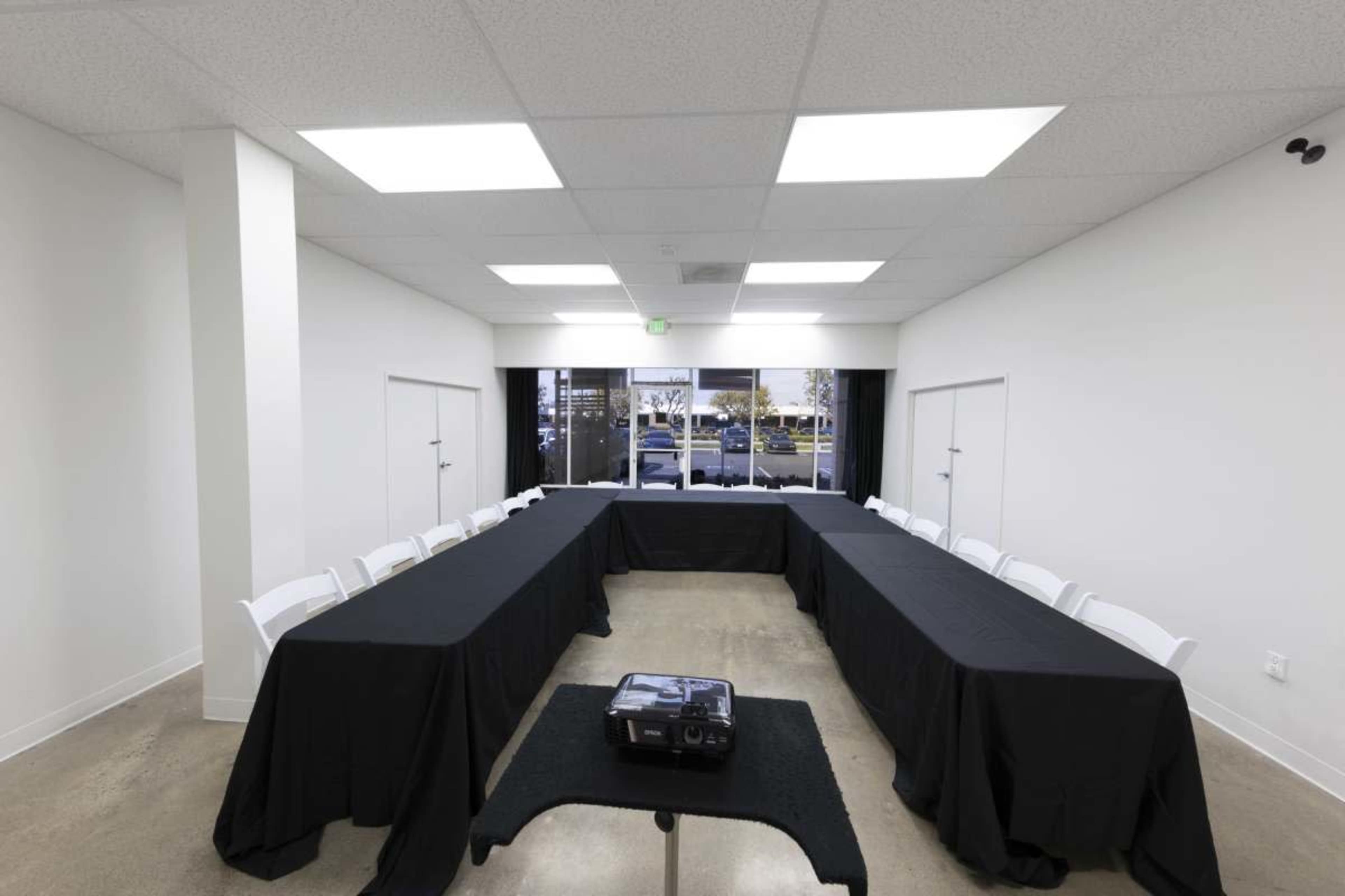 A meeting room features two long tables arranged in a U-shape, covered with black tablecloths, and equipped with a projector at the center, with large windows providing natural light.