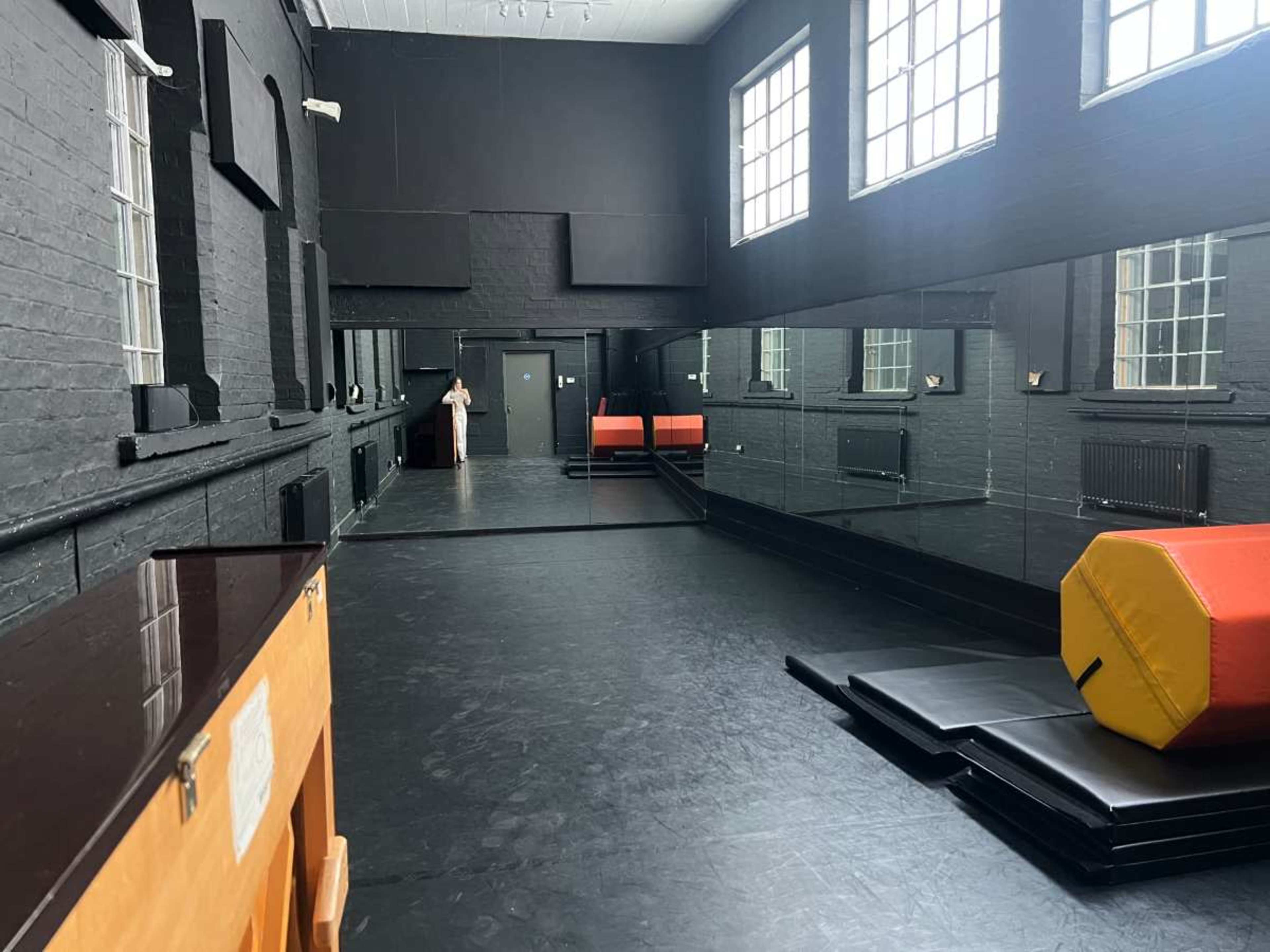 The image shows a spacious, dark-colored gym or studio with large windows, a mirrored wall, and various gymnastic mats and equipment placed along the floor.