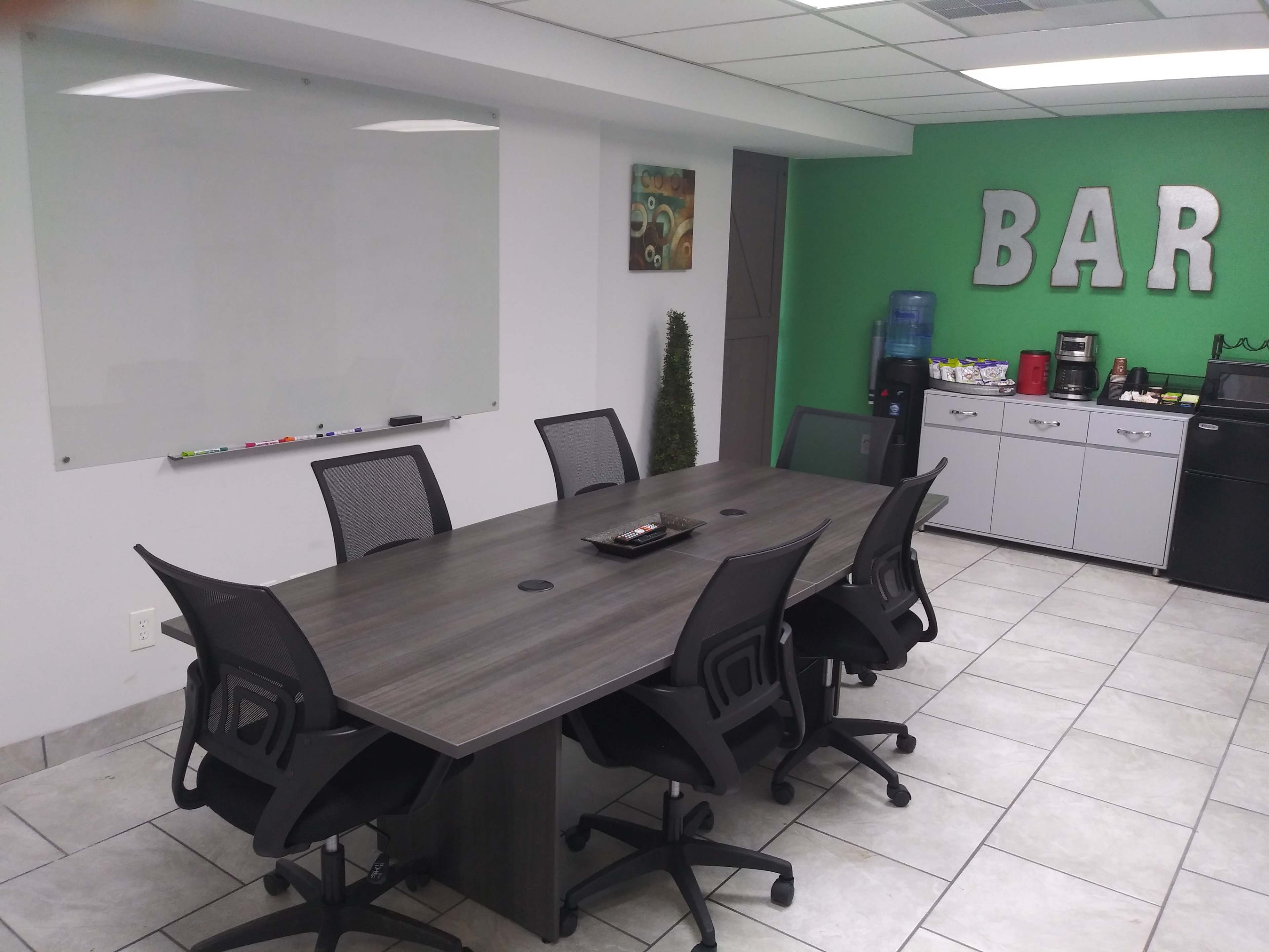 A modern conference room with a rectangular table surrounded by six chairs, featuring a whiteboard, a water cooler, and a kitchenette in the background.