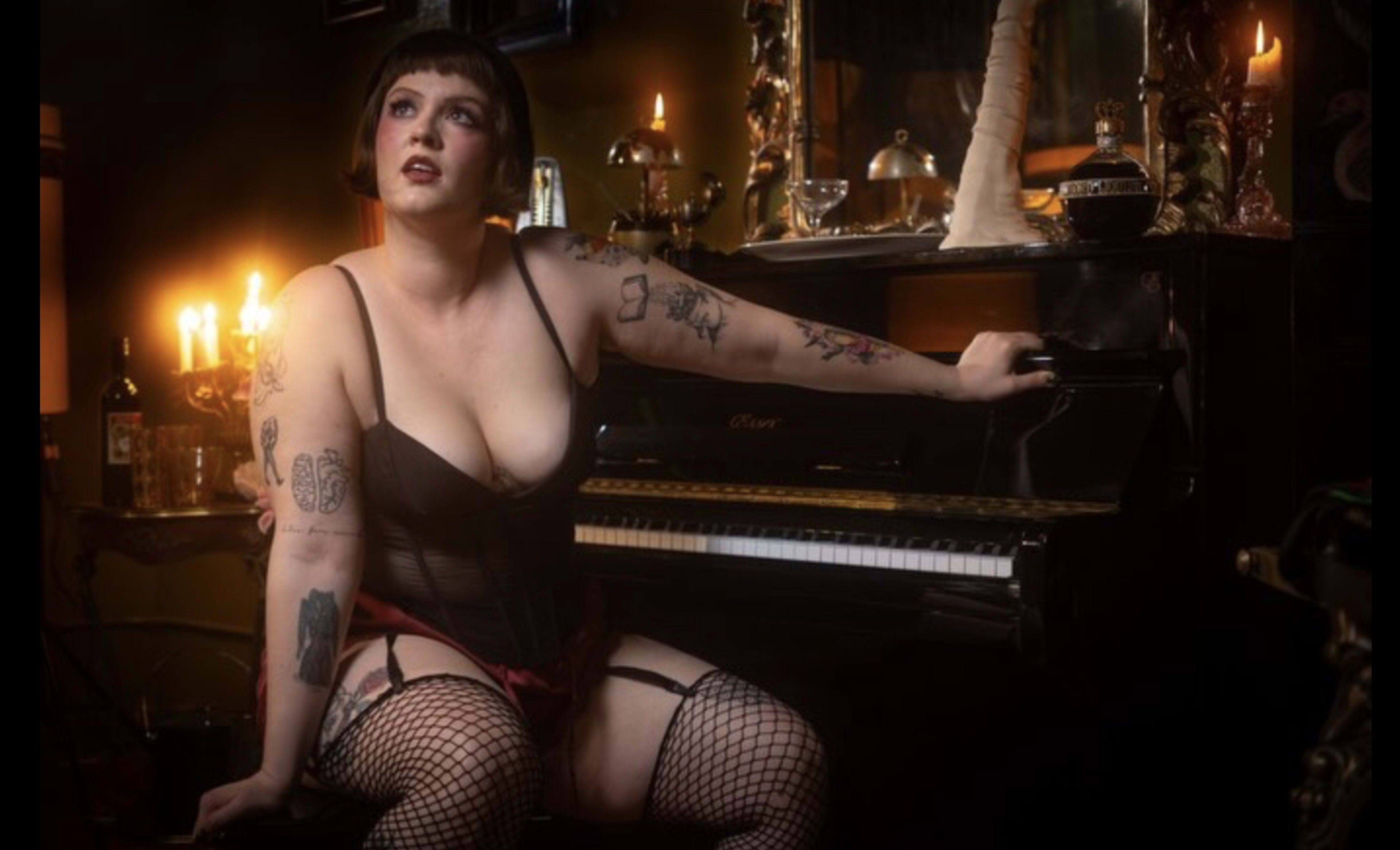 A person with tattoos sits on a piano bench, wearing a black corset and fishnet stockings, illuminated by candlelight in a dimly lit room.