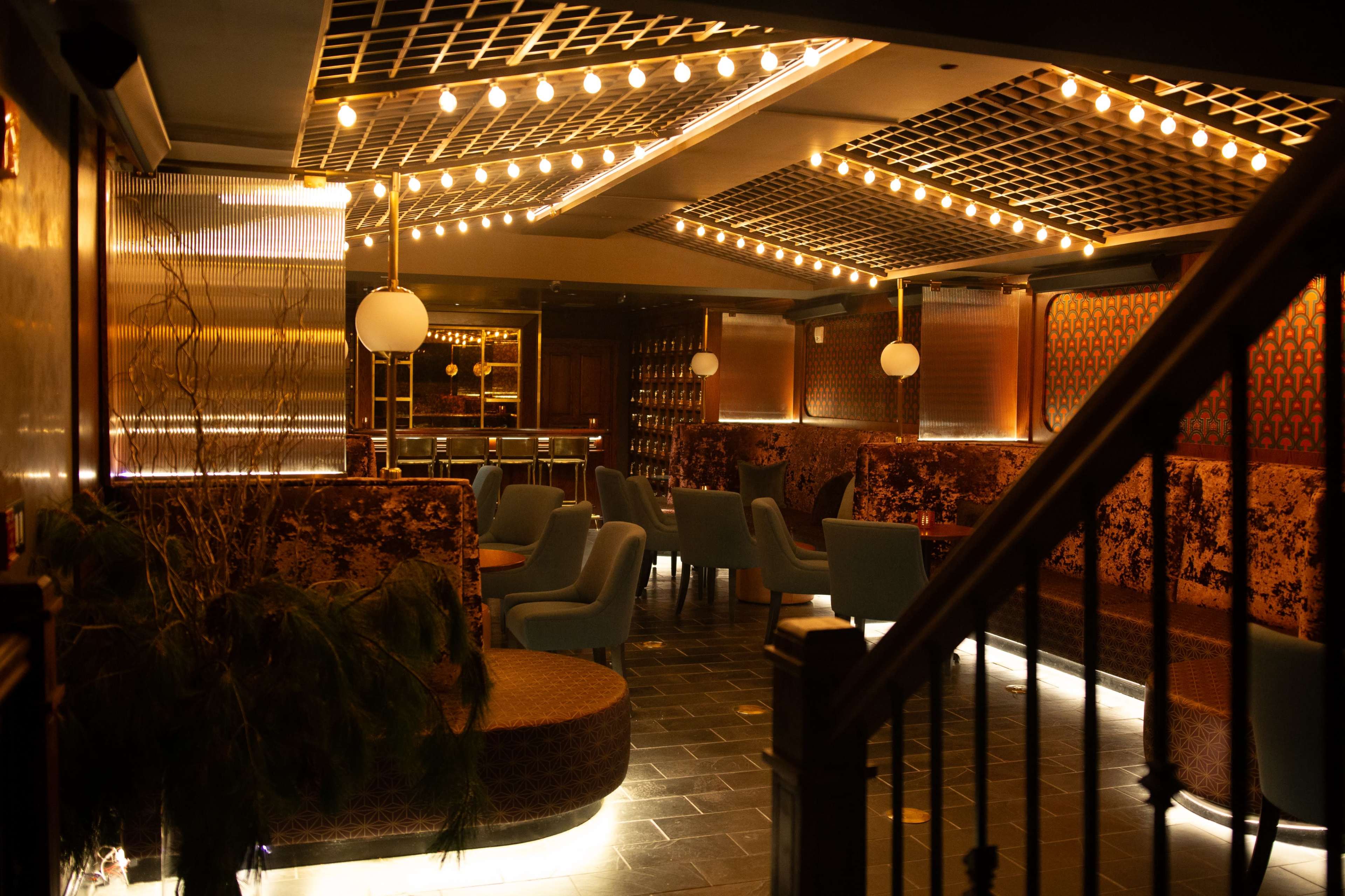 The image depicts a dimly lit lounge or bar area with patterned seating, decorative lighting, and a modern ambiance.