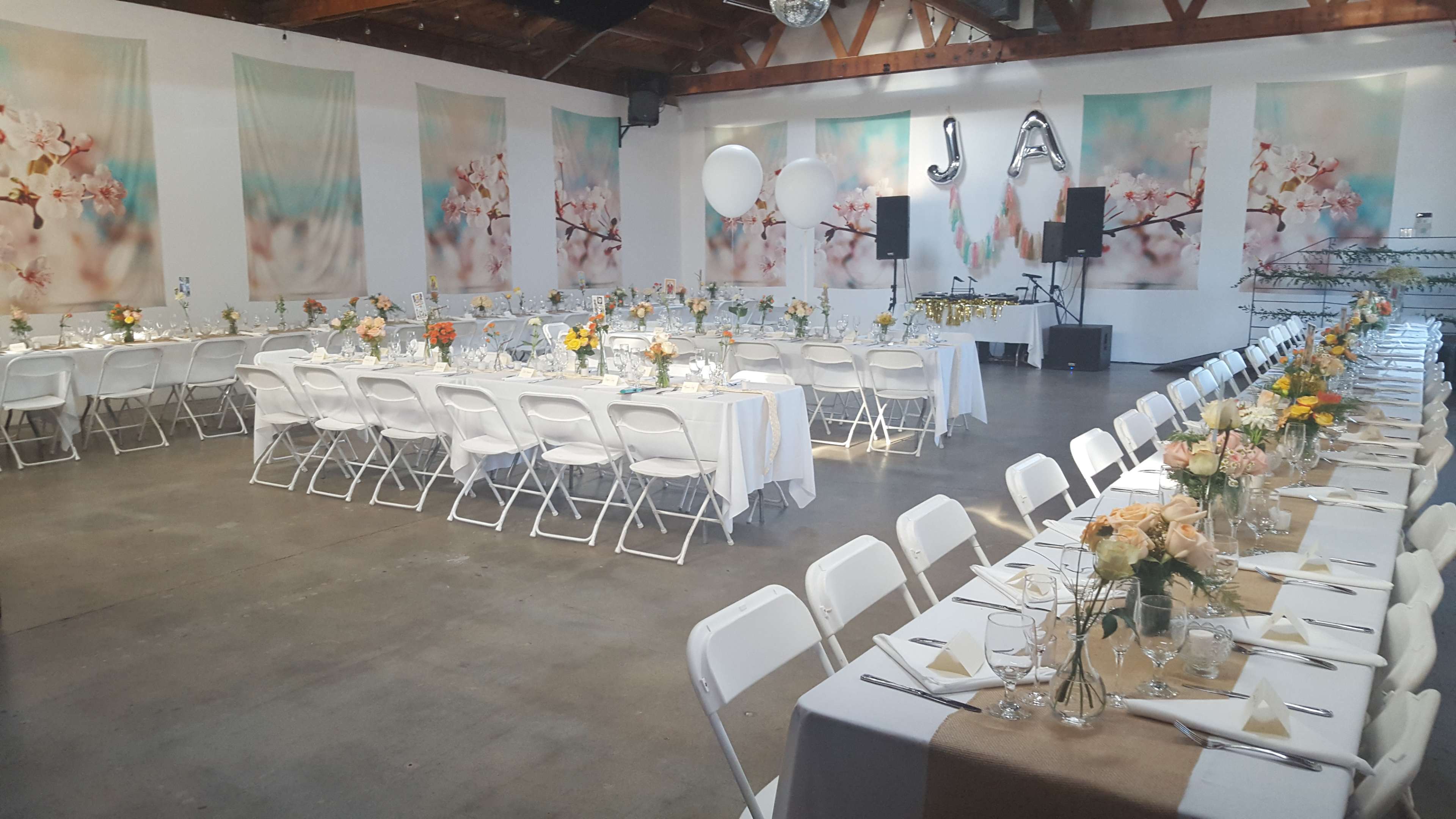 A large indoor event space is set up for a celebration, featuring long tables adorned with floral centerpieces and a backdrop of soft, cherry blossom-themed decorations.