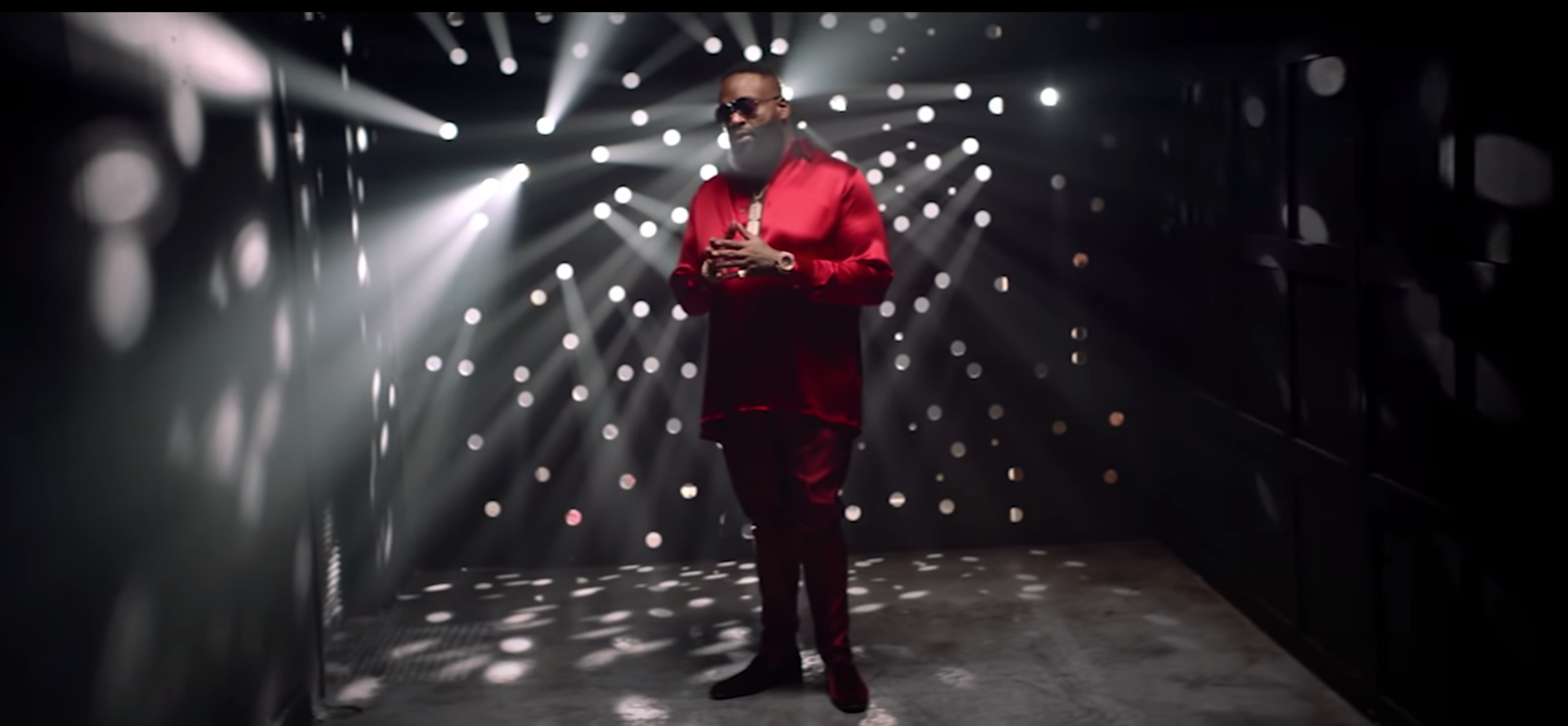 A man dressed in a red outfit stands in a dimly lit space with beams of light and reflections creating a patterned backdrop.