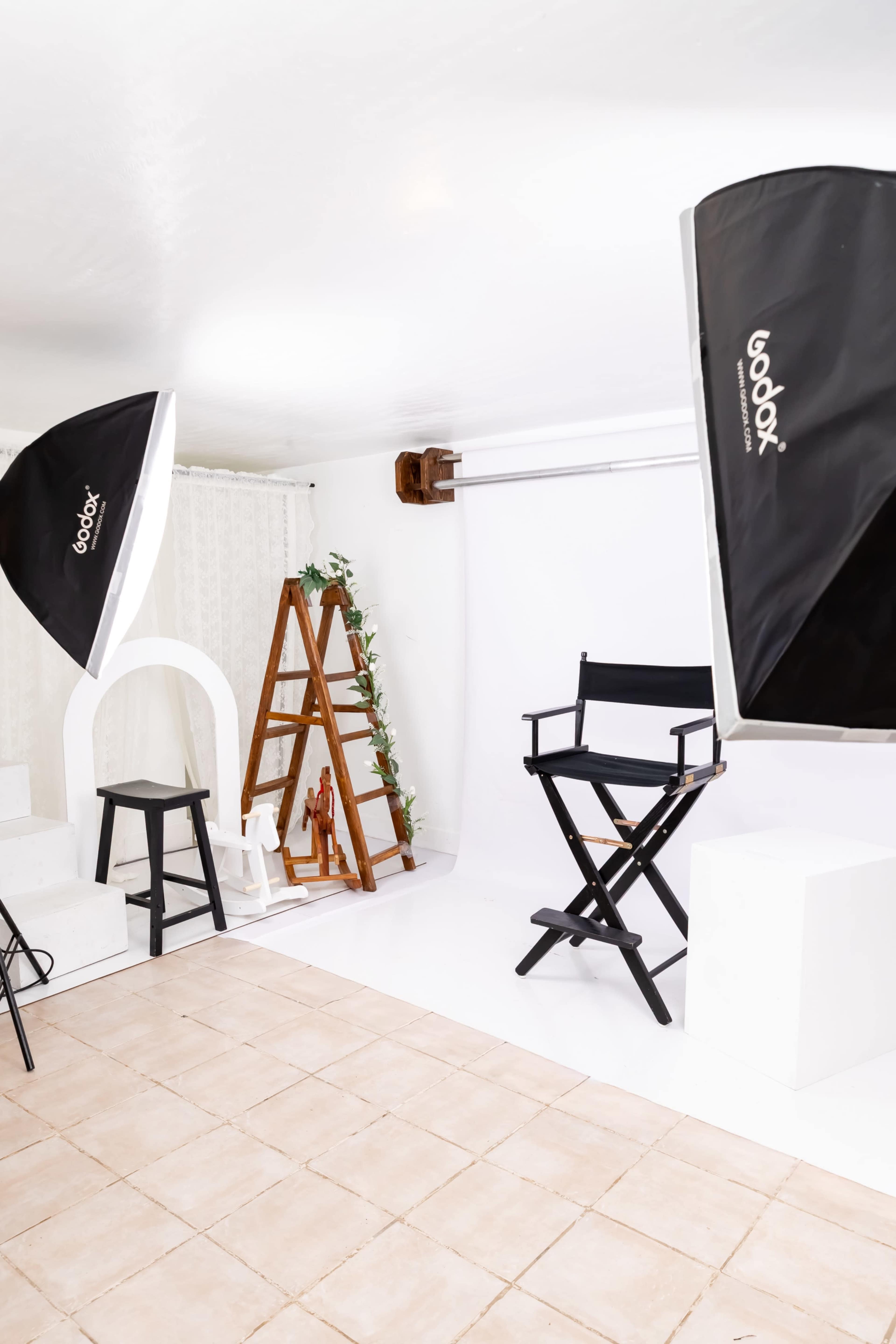 The image shows a photography studio with two softbox lights, a director's chair, a wooden ladder with greenery, and a plain backdrop.