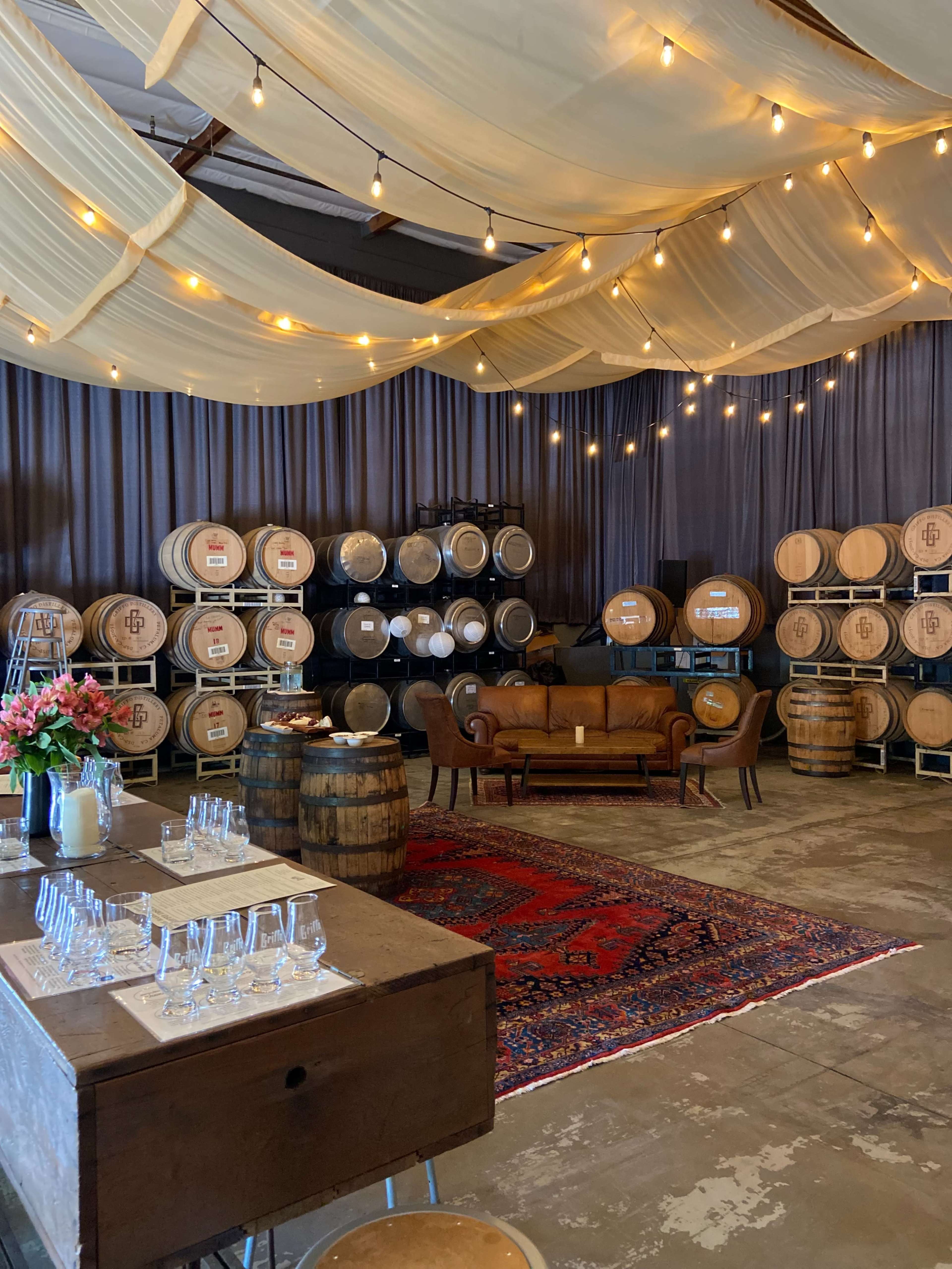 A spacious room features wooden barrels stacked against a wall, a rustic couch, and a decorated table with glassware, all under string lights and draped fabric.