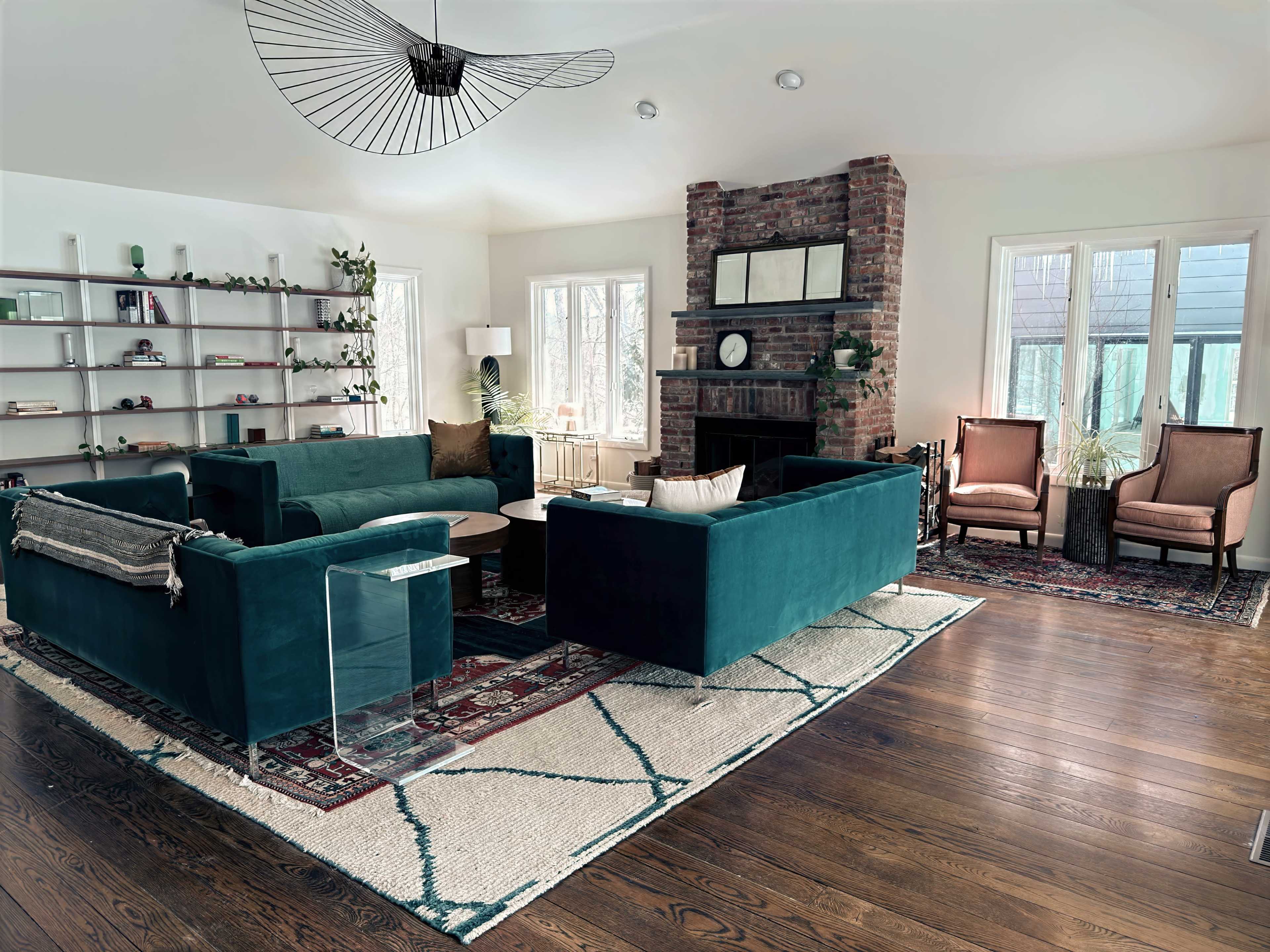 The living room features a pair of teal sectional sofas surrounding a round coffee table, with a brick fireplace and large windows in the background.