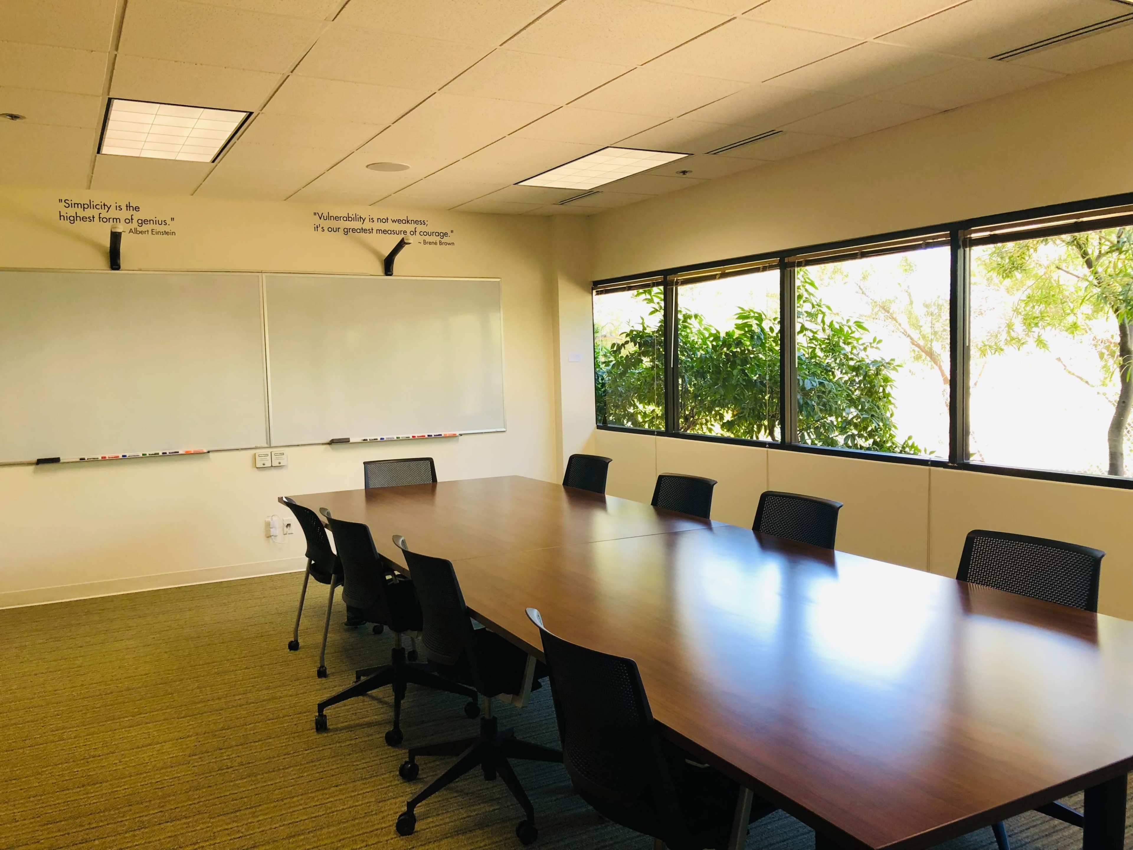 A meeting room features a long wooden table surrounded by black chairs, with large windows overlooking greenery and two whiteboards on the wall.