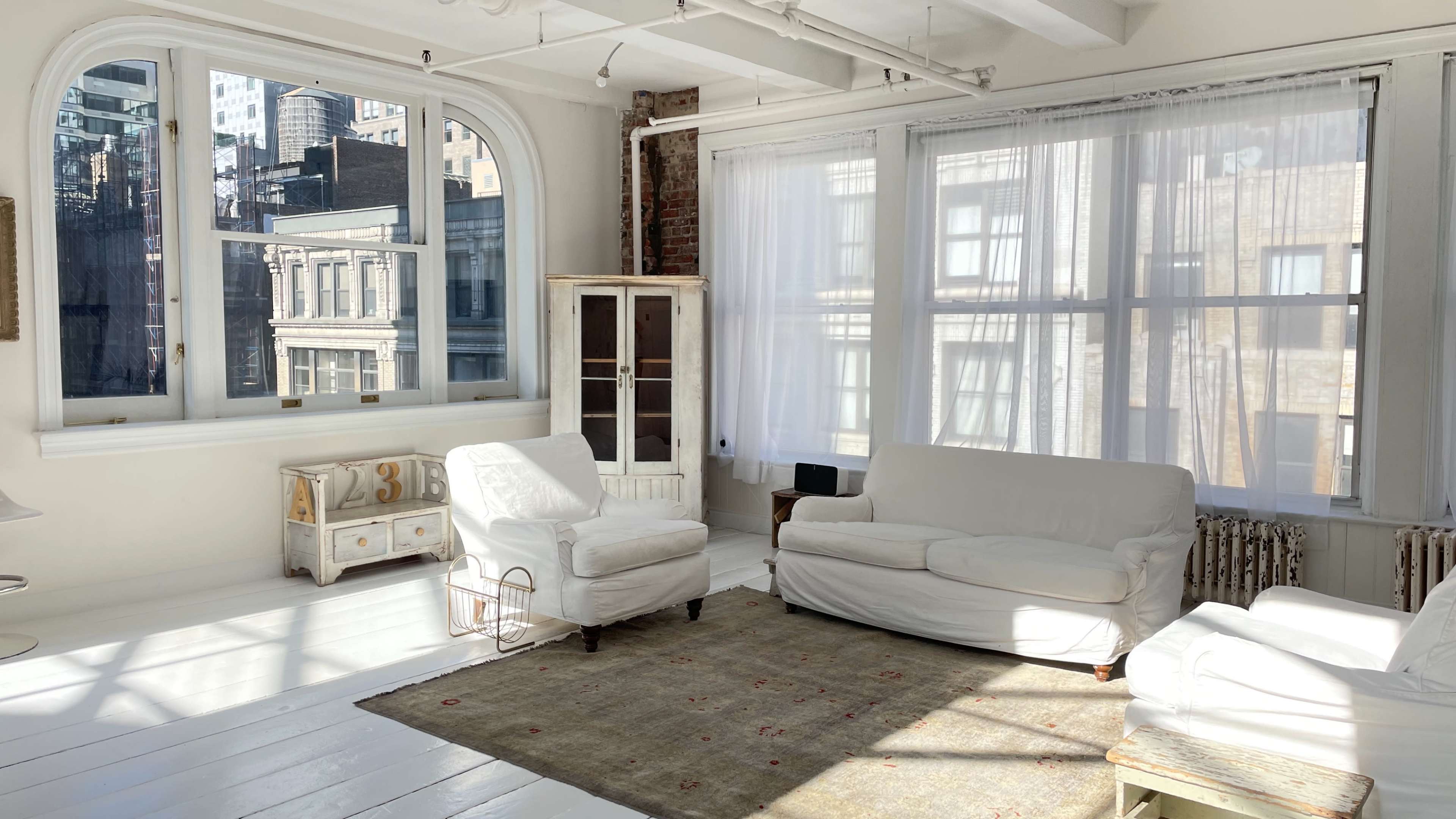 The image shows a bright, modern living room with white furniture, large windows, and a light rug on a wooden floor.