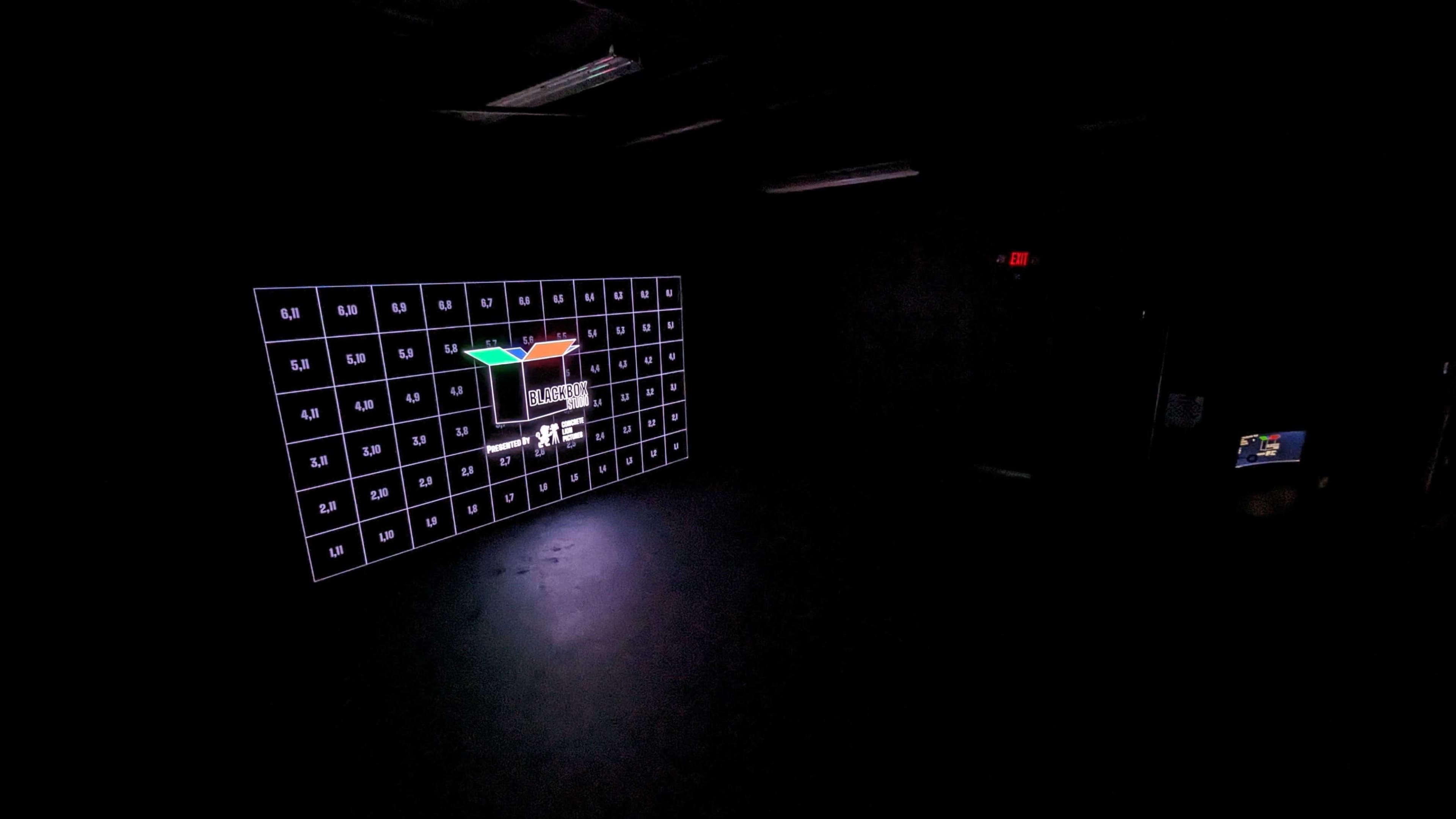A dimly lit room featuring a large wall display with a grid of glowing squares and a colorful graphical element in the center.