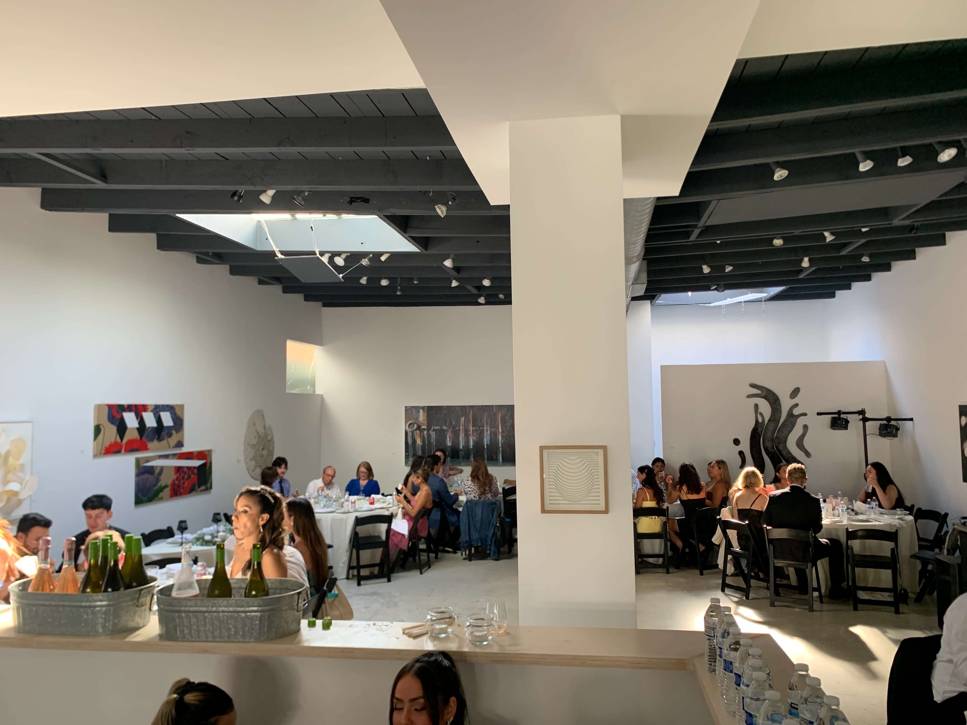 A group of diners sits at tables in a well-lit restaurant featuring white walls and black beams, with artworks displayed throughout the space.