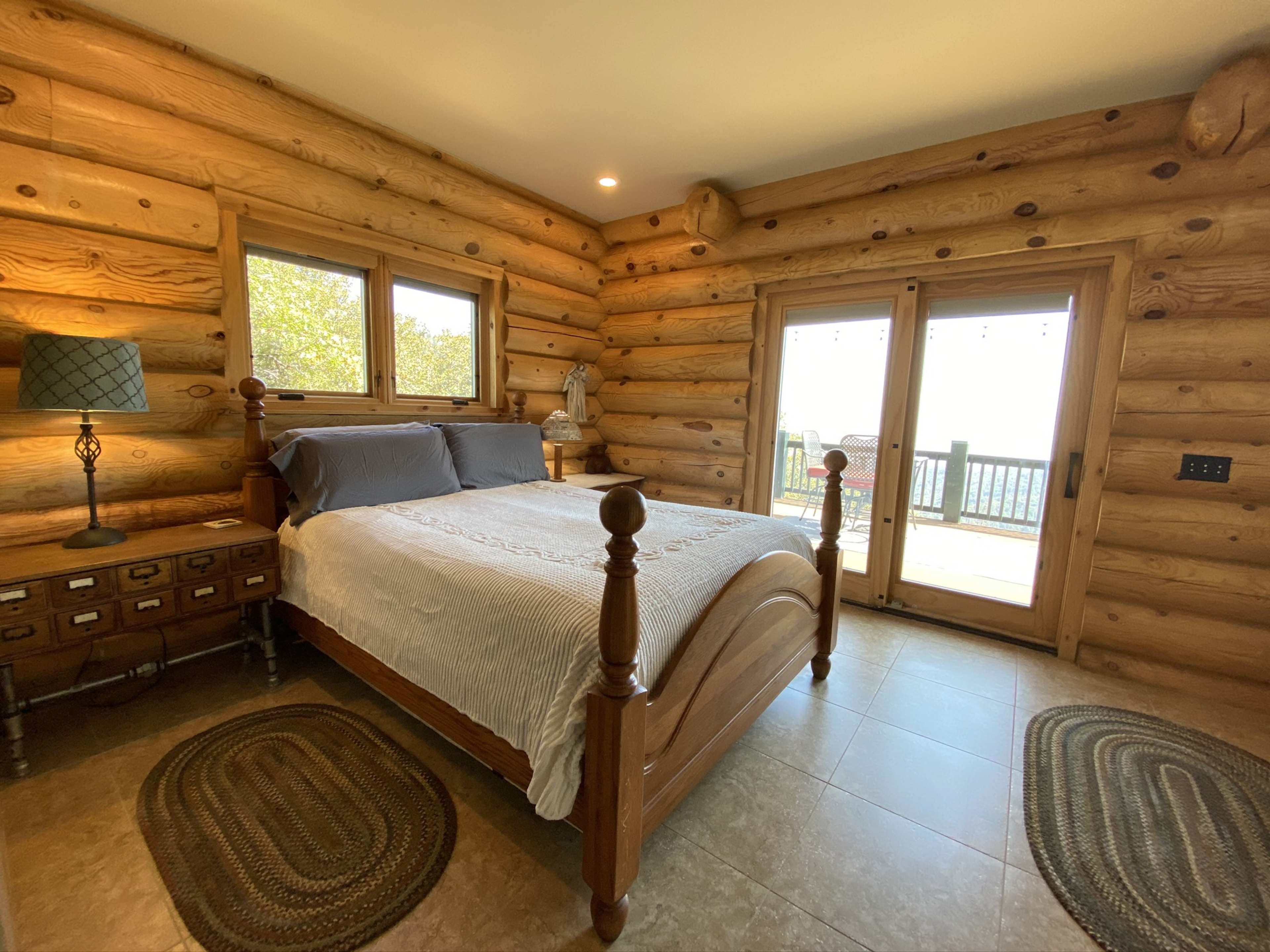 The image shows a log cabin bedroom featuring a wooden bed, side tables, and large windows with views of a deck outside.
