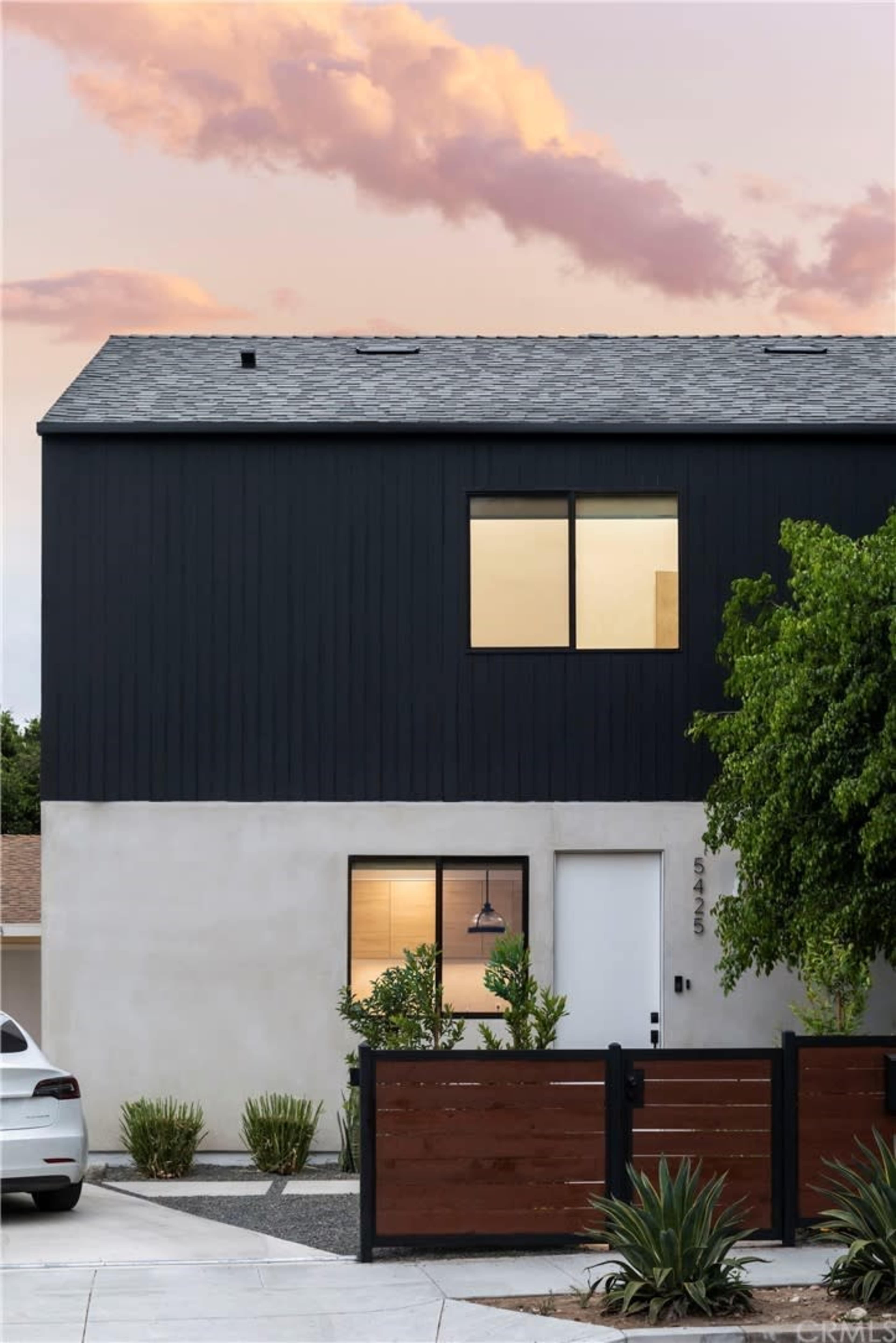 A modern two-story house with dark siding and a light-colored lower section, surrounded by landscaped greenery and a driveway.