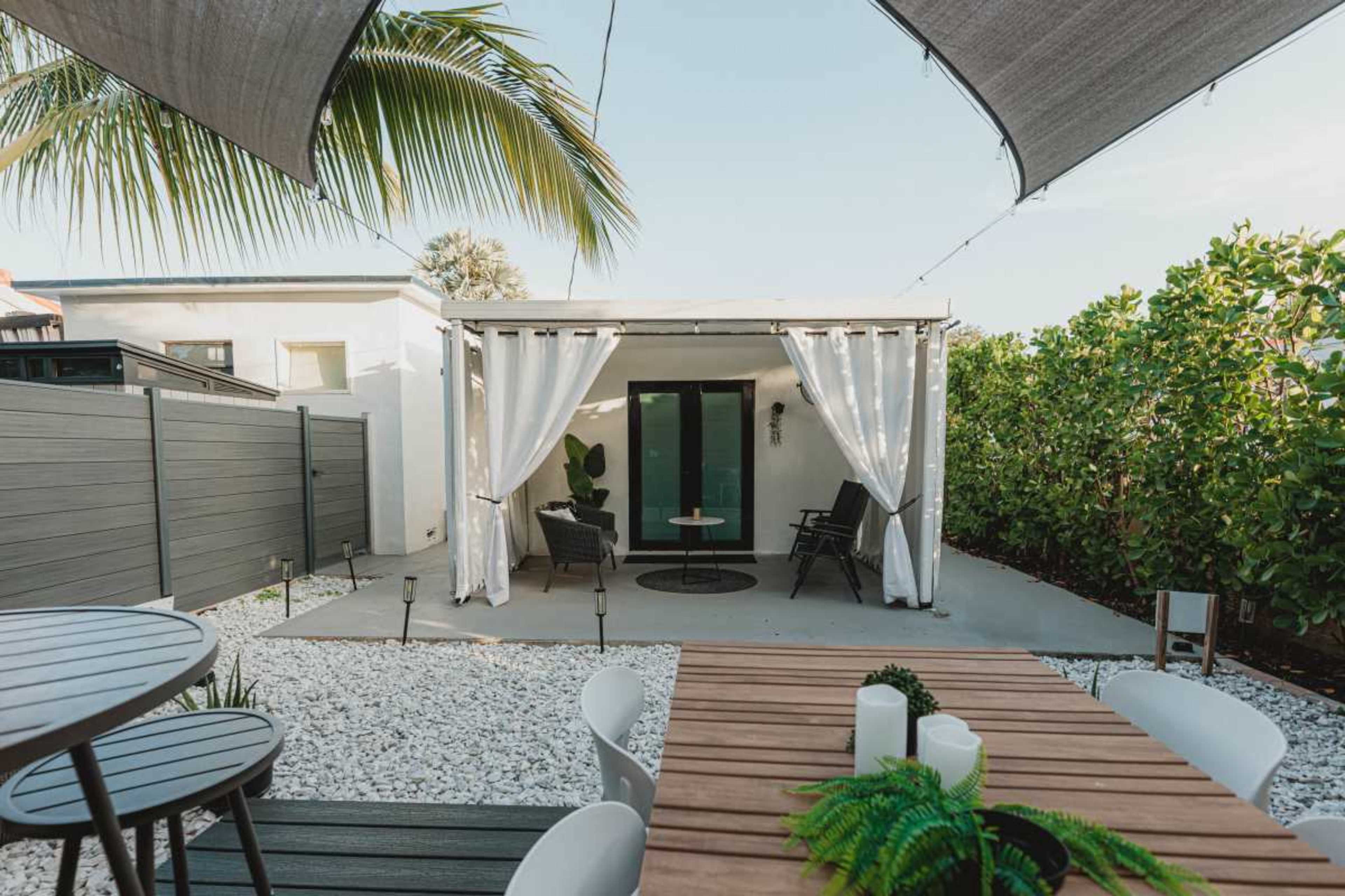 The image shows a patio area with a wooden dining table and chairs, surrounded by white gravel, leading to a covered seating area with curtains and two chairs.