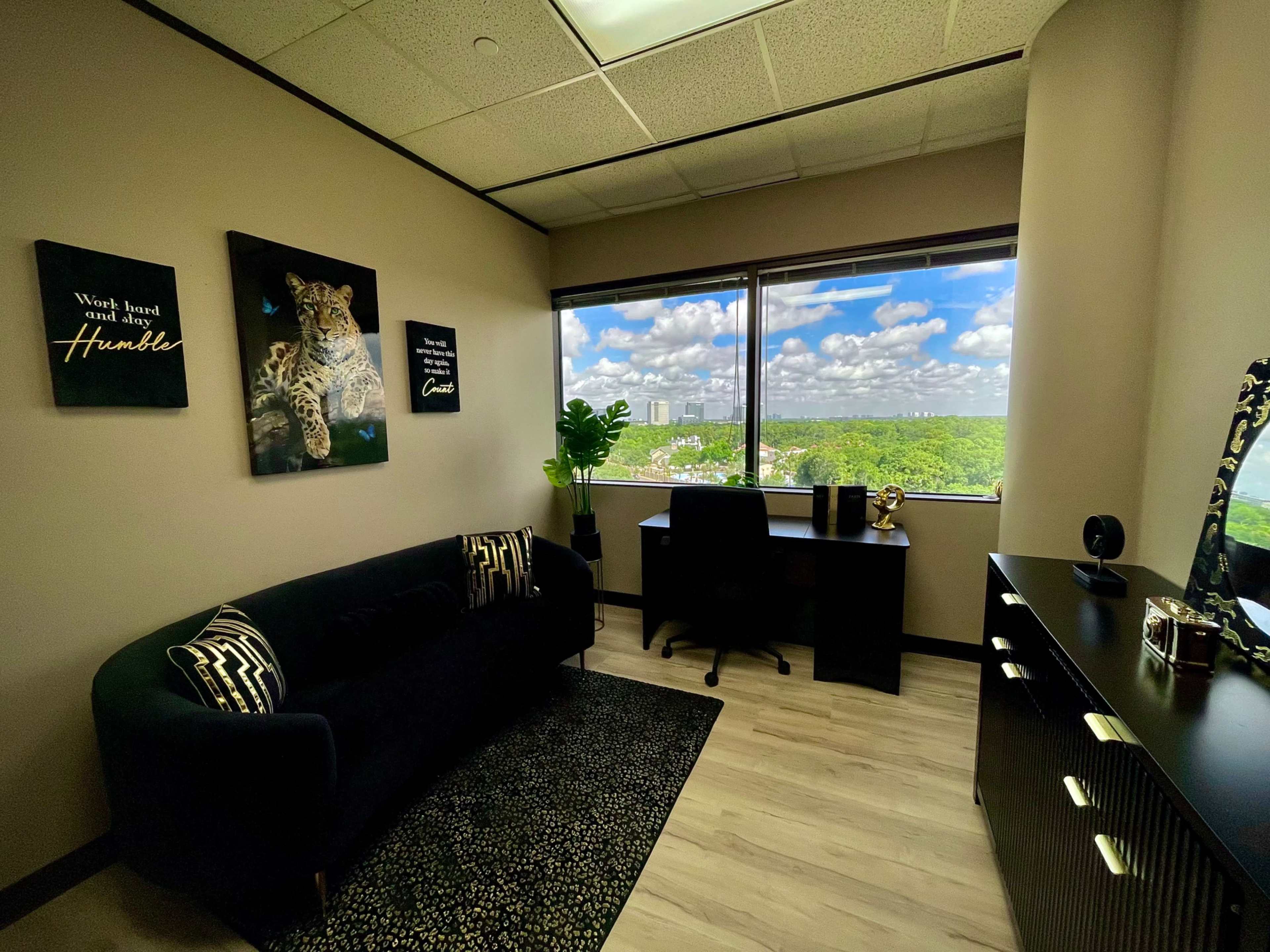 A modern office features a dark couch and decorative artwork on the walls, with a large window that offers a view of a lush green landscape and sky filled with clouds.