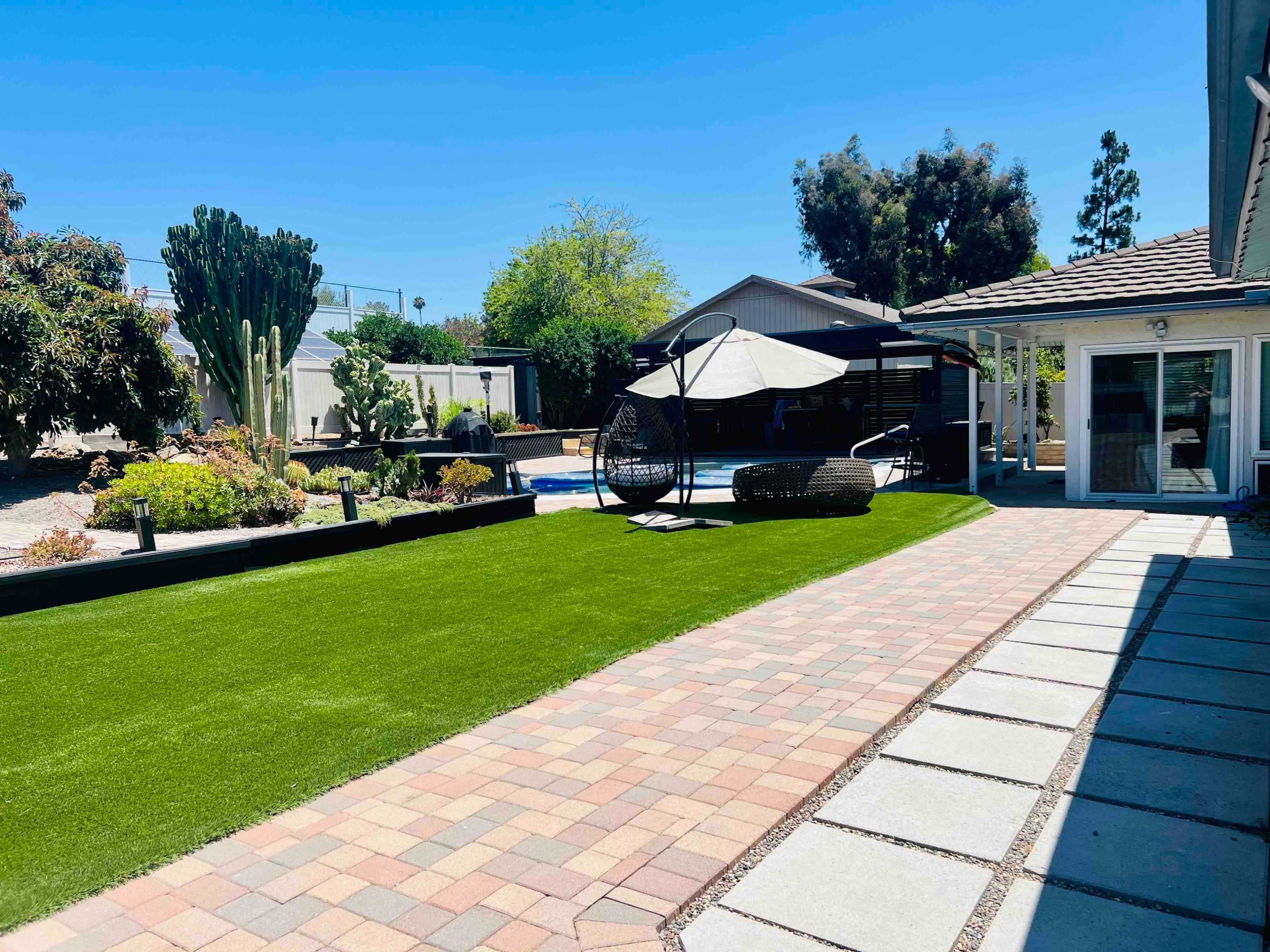 The image shows a landscaped backyard featuring artificial grass, a stone pathway, a hanging chair, and a swimming pool surrounded by various plants and trees.