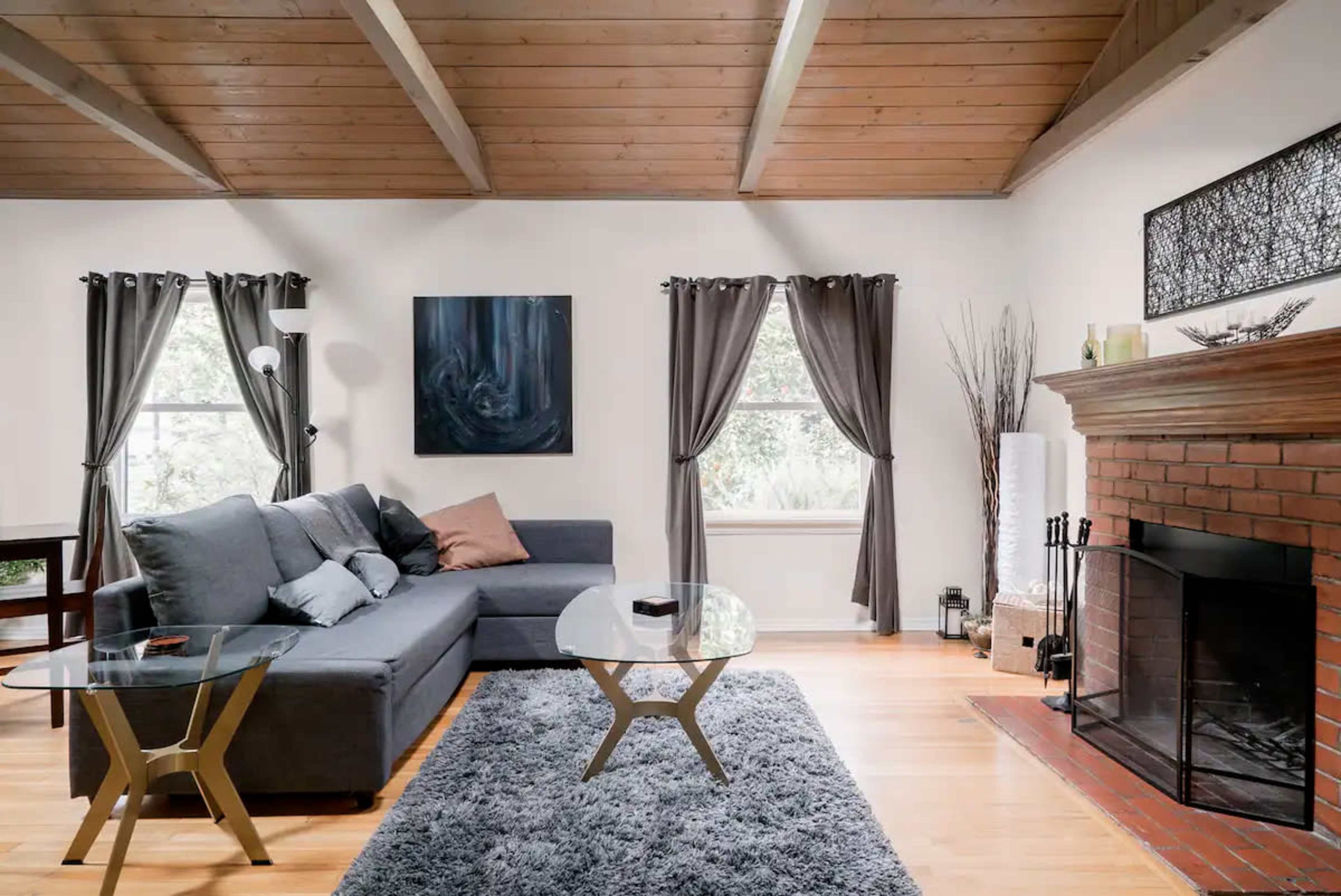 A cozy living room features a gray sectional sofa, two glass-top tables, a brick fireplace, and large windows with dark curtains.