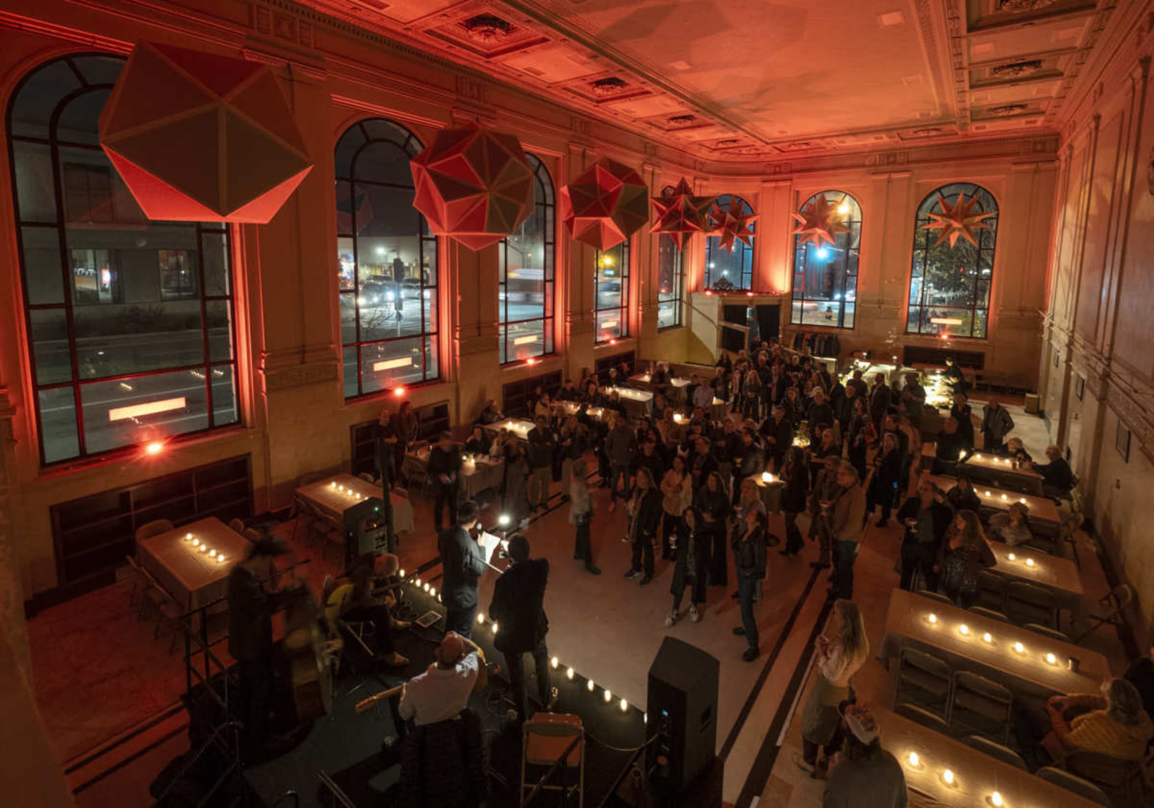 A large hall filled with people enjoying an event, illuminated by red lighting and large decorative shapes hanging from the ceiling.