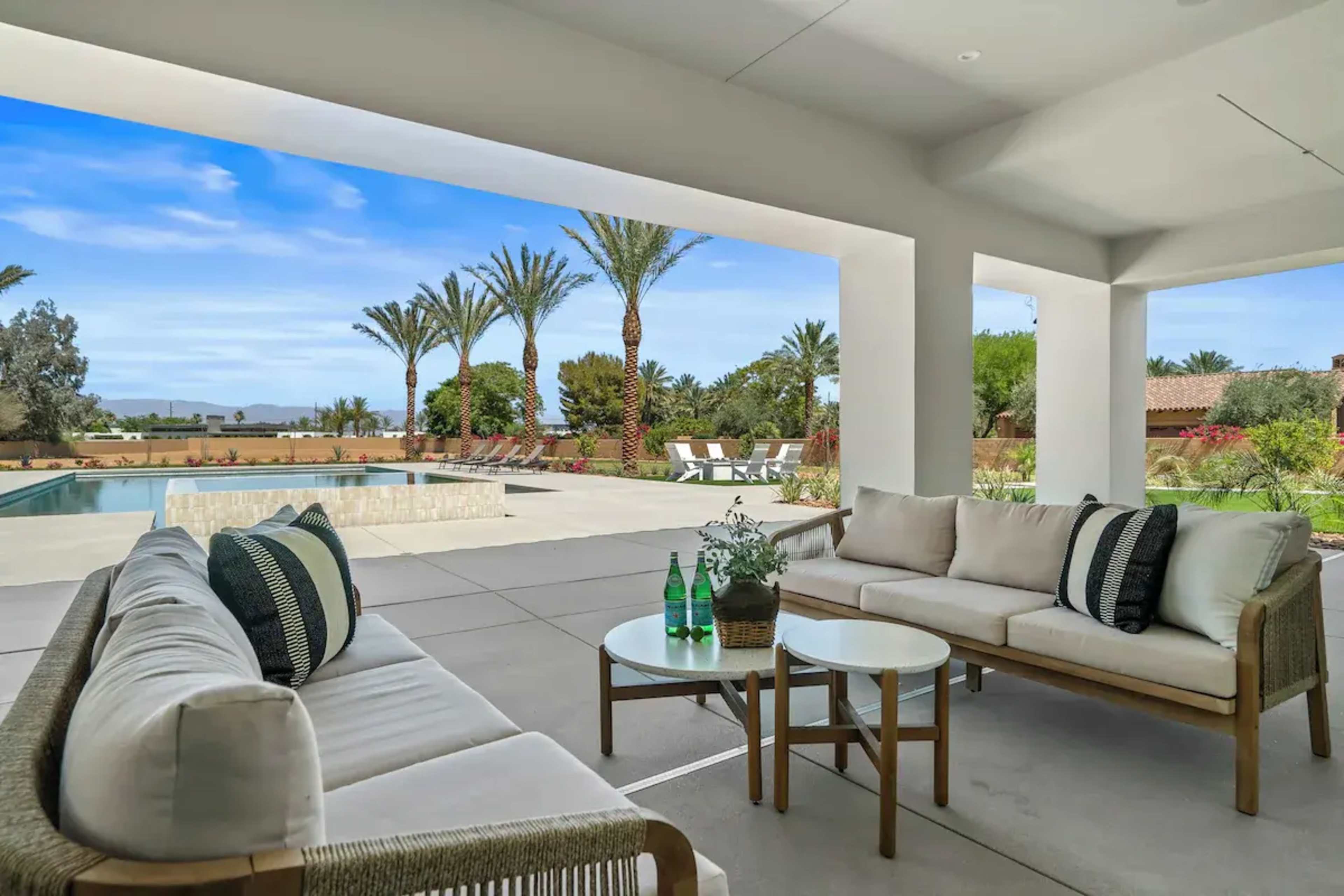 The image shows a modern outdoor patio featuring a seating area with a sofa and coffee table, overlooking a pool and landscaped garden with palm trees.
