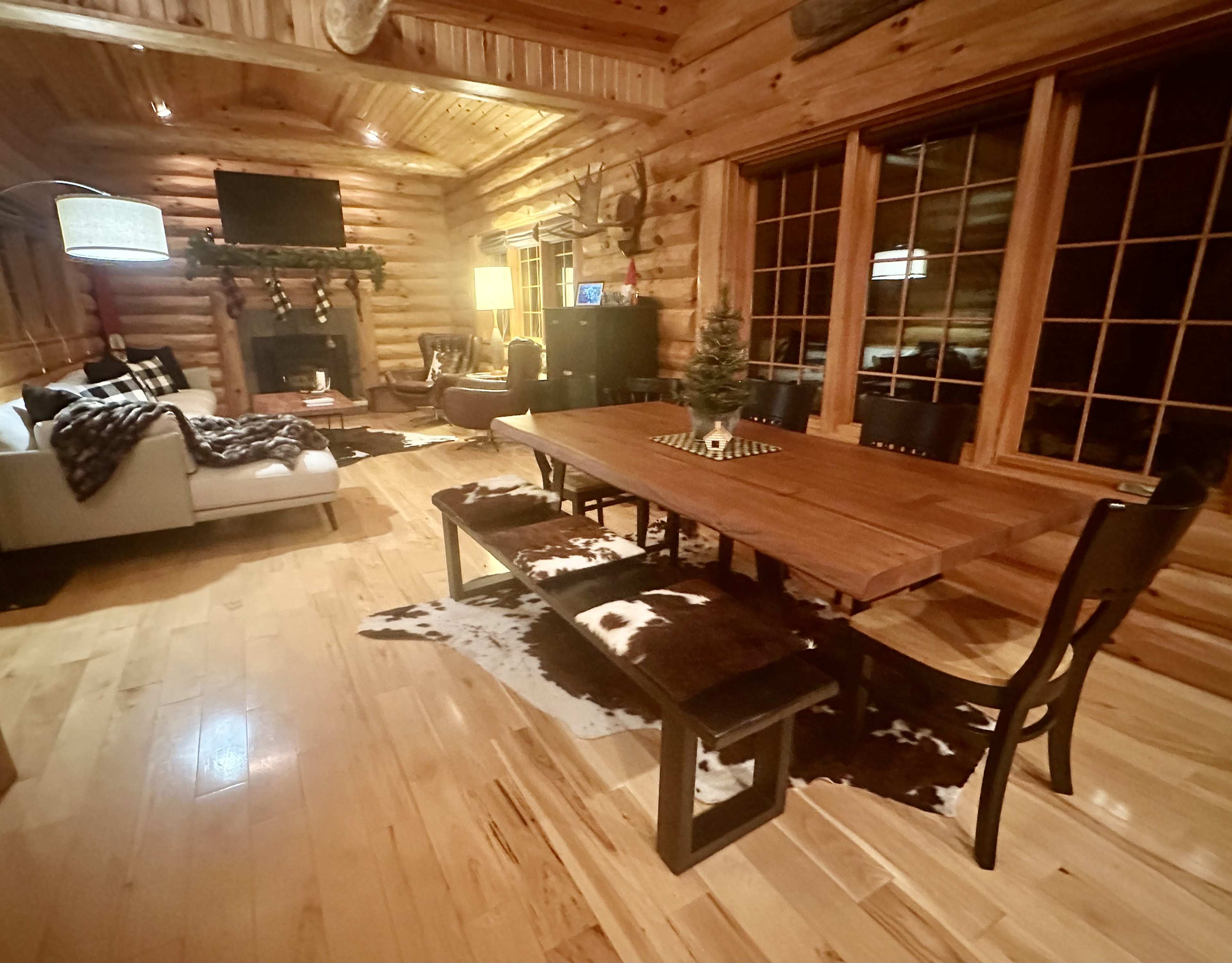 The image shows a rustic log cabin interior with a wooden dining table, a seating area featuring a couch, and a fireplace adorned with decorative elements.