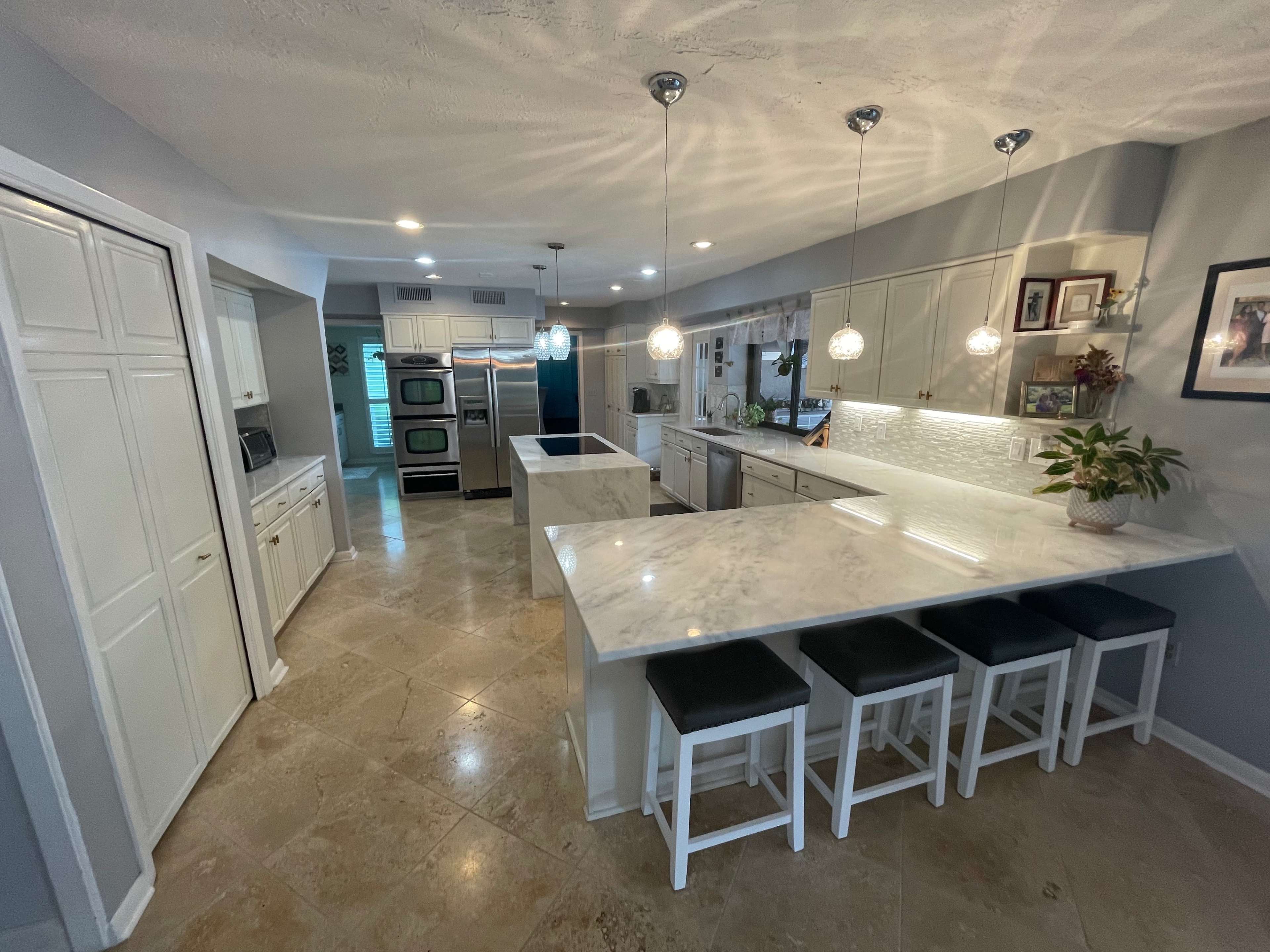 A modern kitchen with a marble countertop, white cabinetry, and pendant lighting over a bar area with four stools.