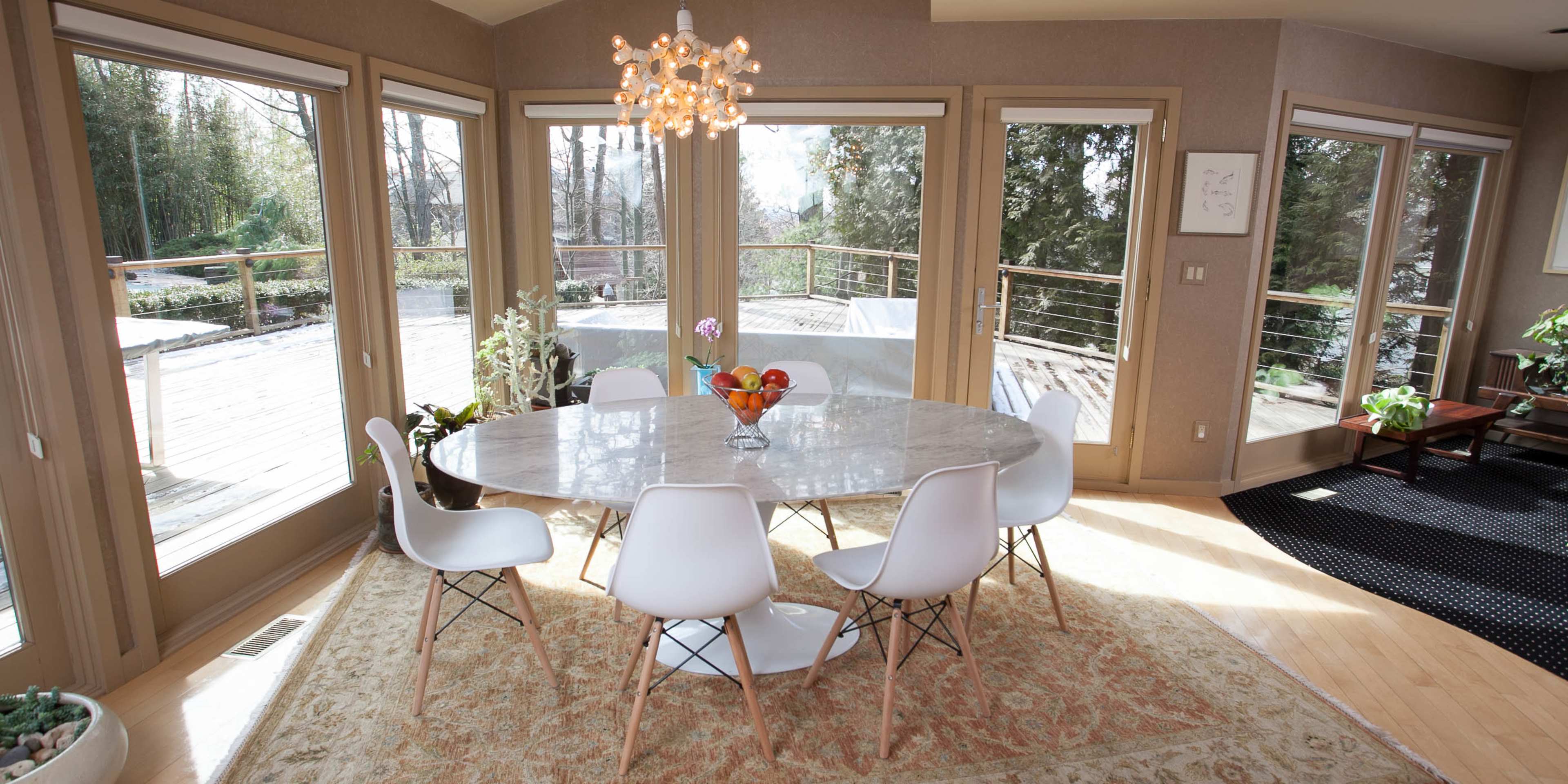 A round marble dining table surrounded by white chairs is set in a sunlit room with large windows overlooking a deck and garden.