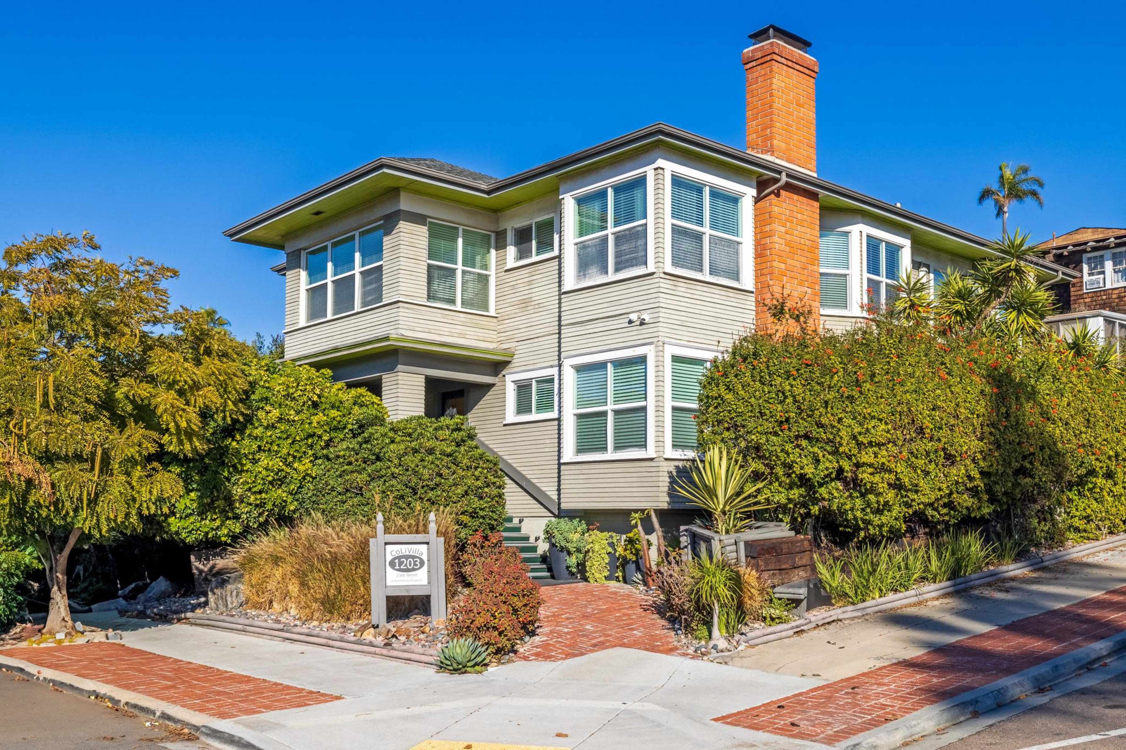 A three-story house with a brick chimney is surrounded by dense greenery and landscaping at a street corner.